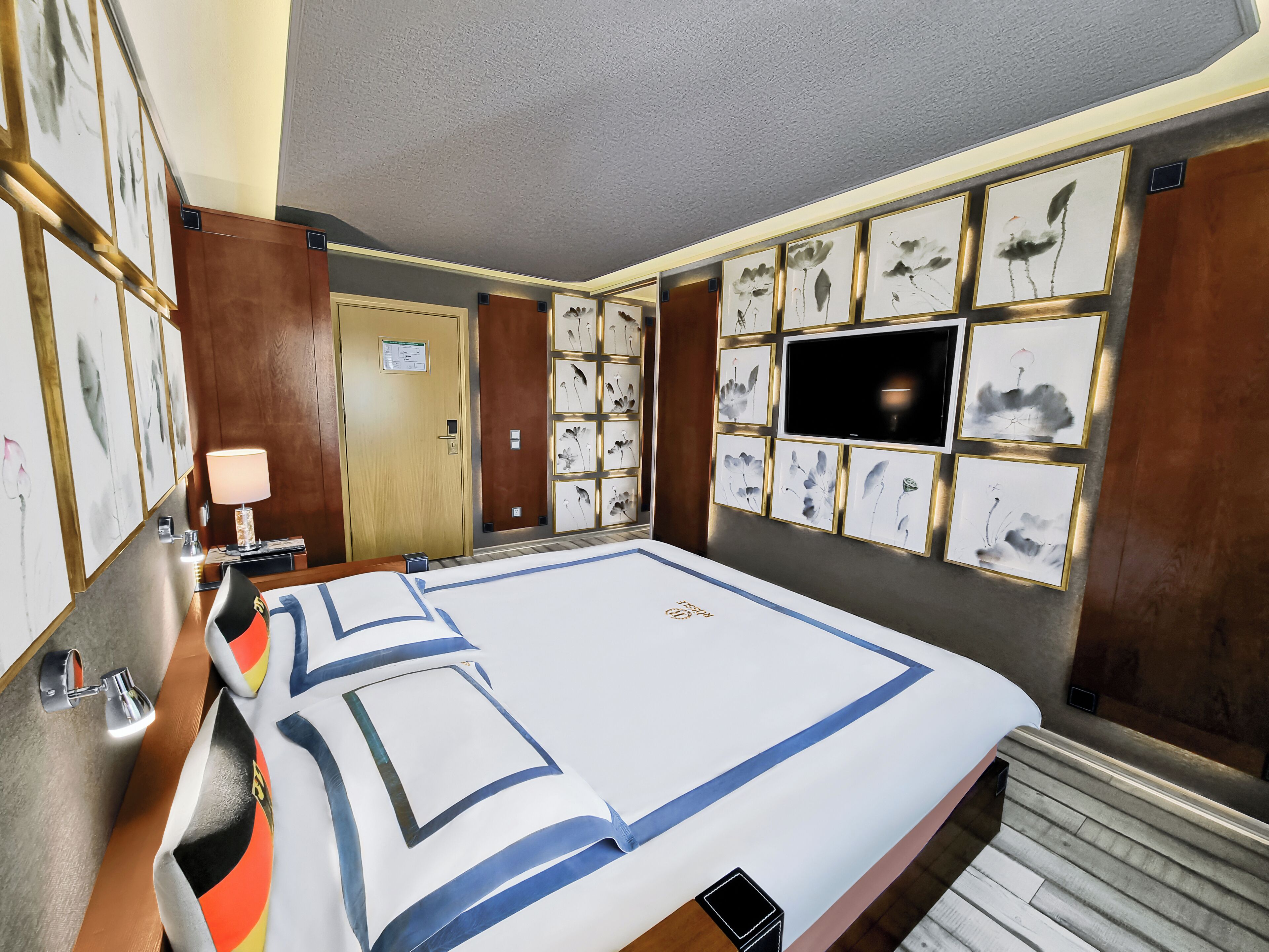 A hotel room with a large bed, a wooden door, a wall-mounted TV, and framed artwork.