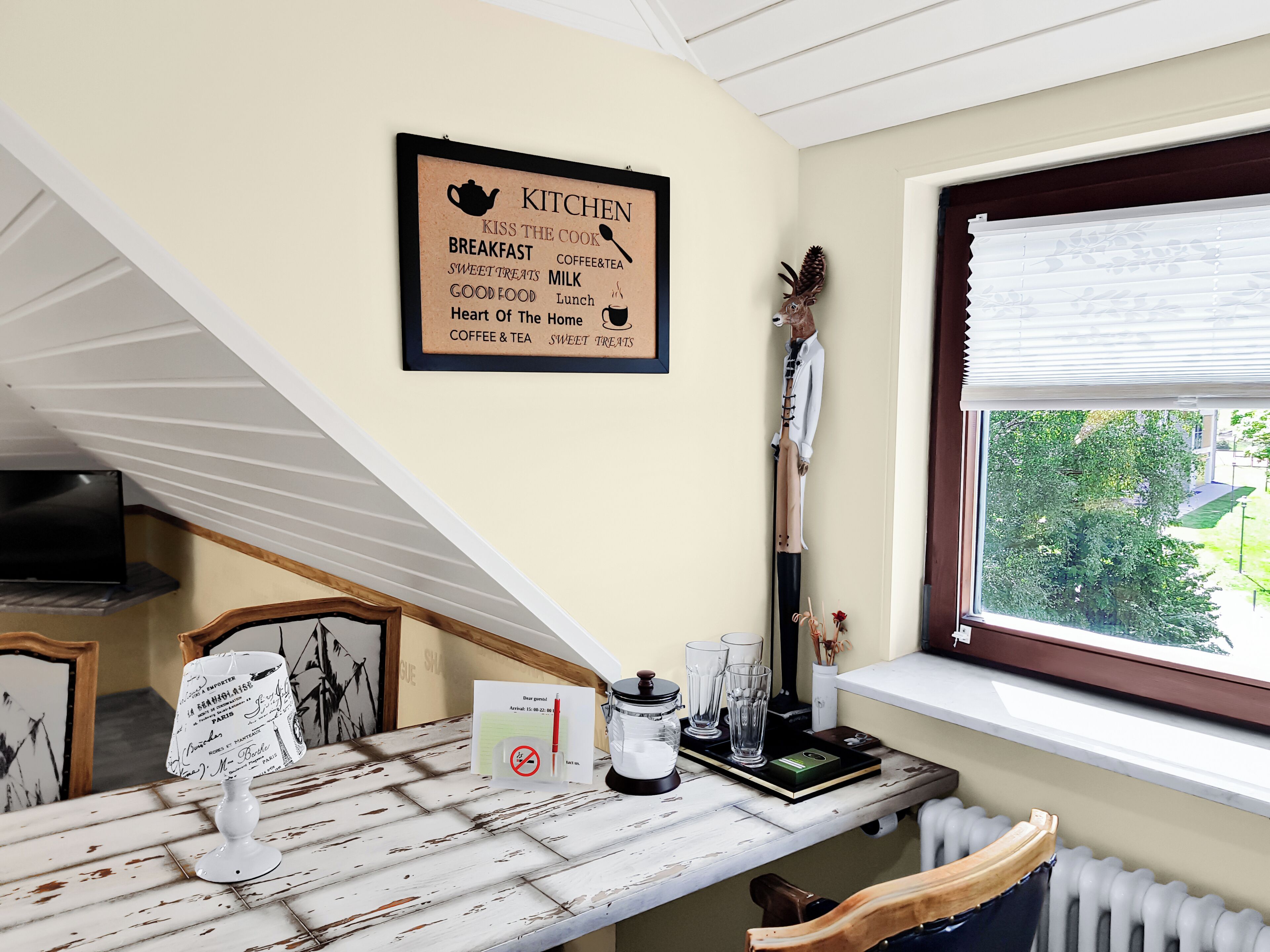 A kitchen with a framed sign listing breakfast items, a window with blinds, a radiator, and a desk with a lamp and a notepad.