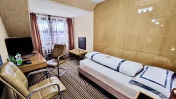 Ein Hotelzimmer mit einem Bett, einem Schreibtisch mit Computer, einem Sessel, einem Fernseher und einem Fenster mit Vorhängen.
