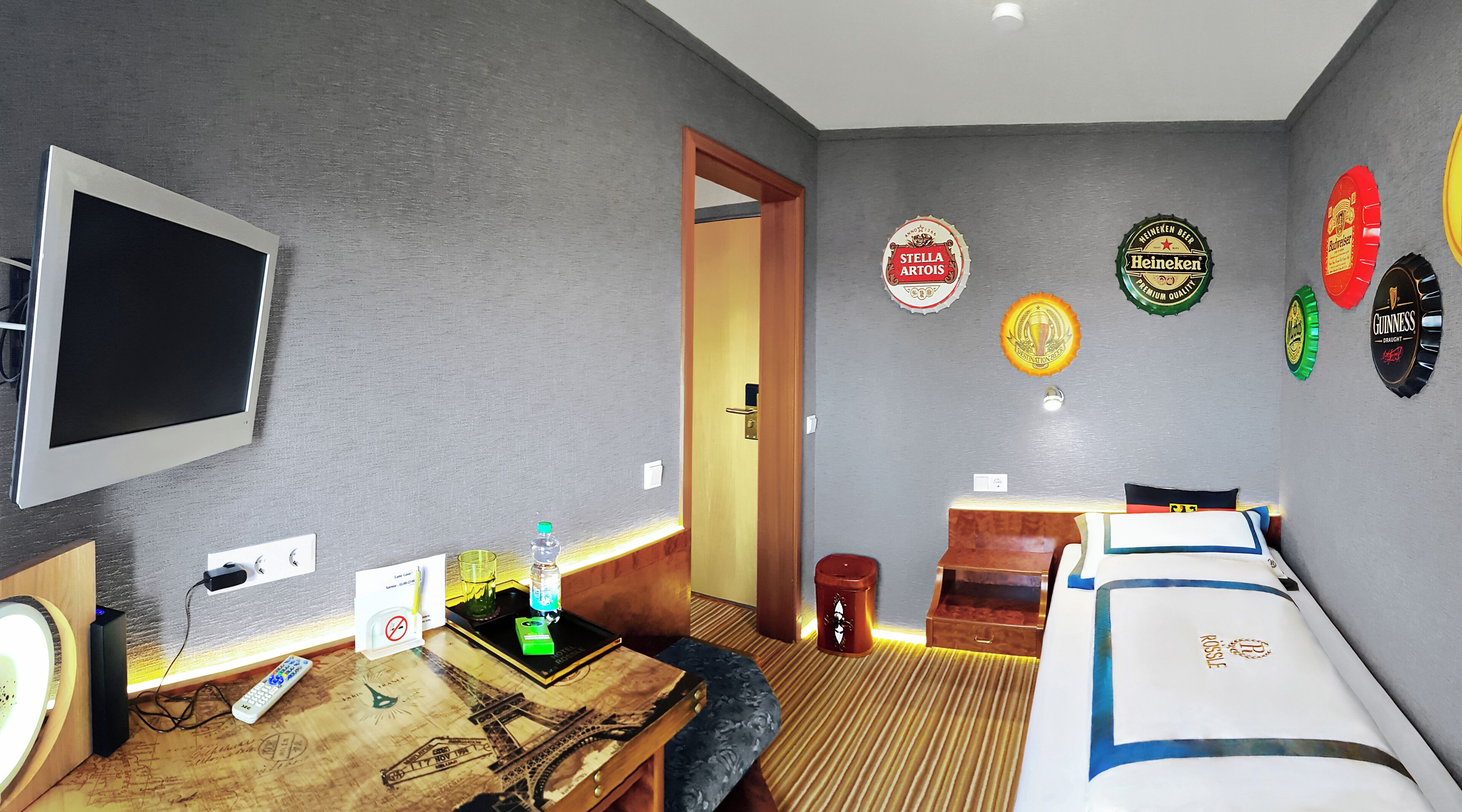 A hotel room with a bed, a desk with a computer, a TV, and wall decorations including beer bottle labels.