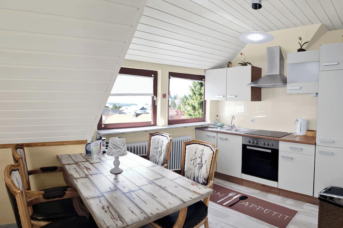 Una cocina compacta con armarios blancos, una mesa de comedor con sillas, un ventanal con persianas y vistas al exterior.