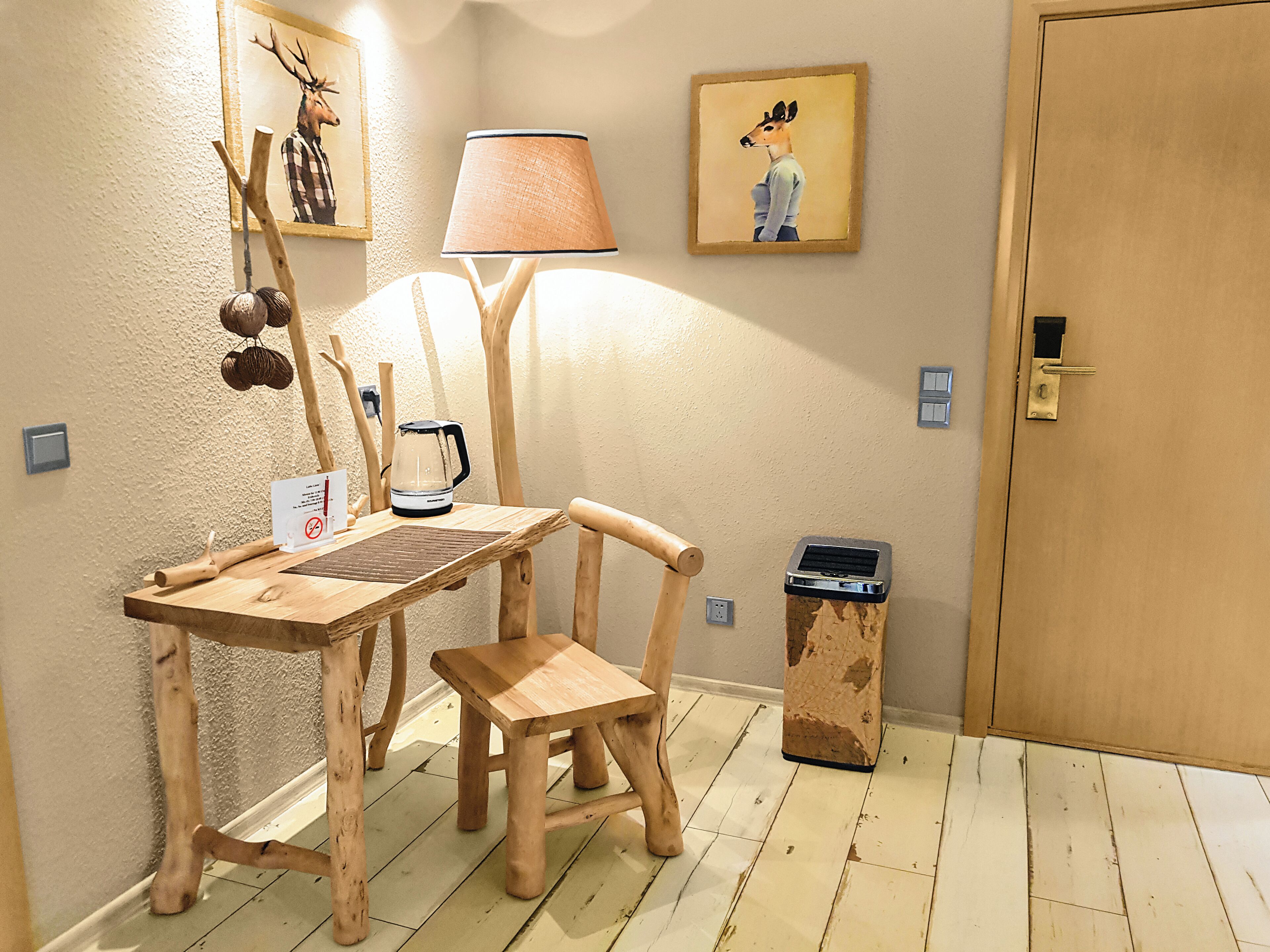 A wooden table with a kettle, a lamp, and a chair in a room with a door and a trash bin.