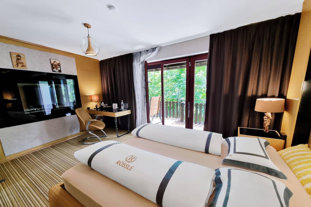 Habitación de hotel con cama, escritorio, silla, televisor y balcón con cortinas.