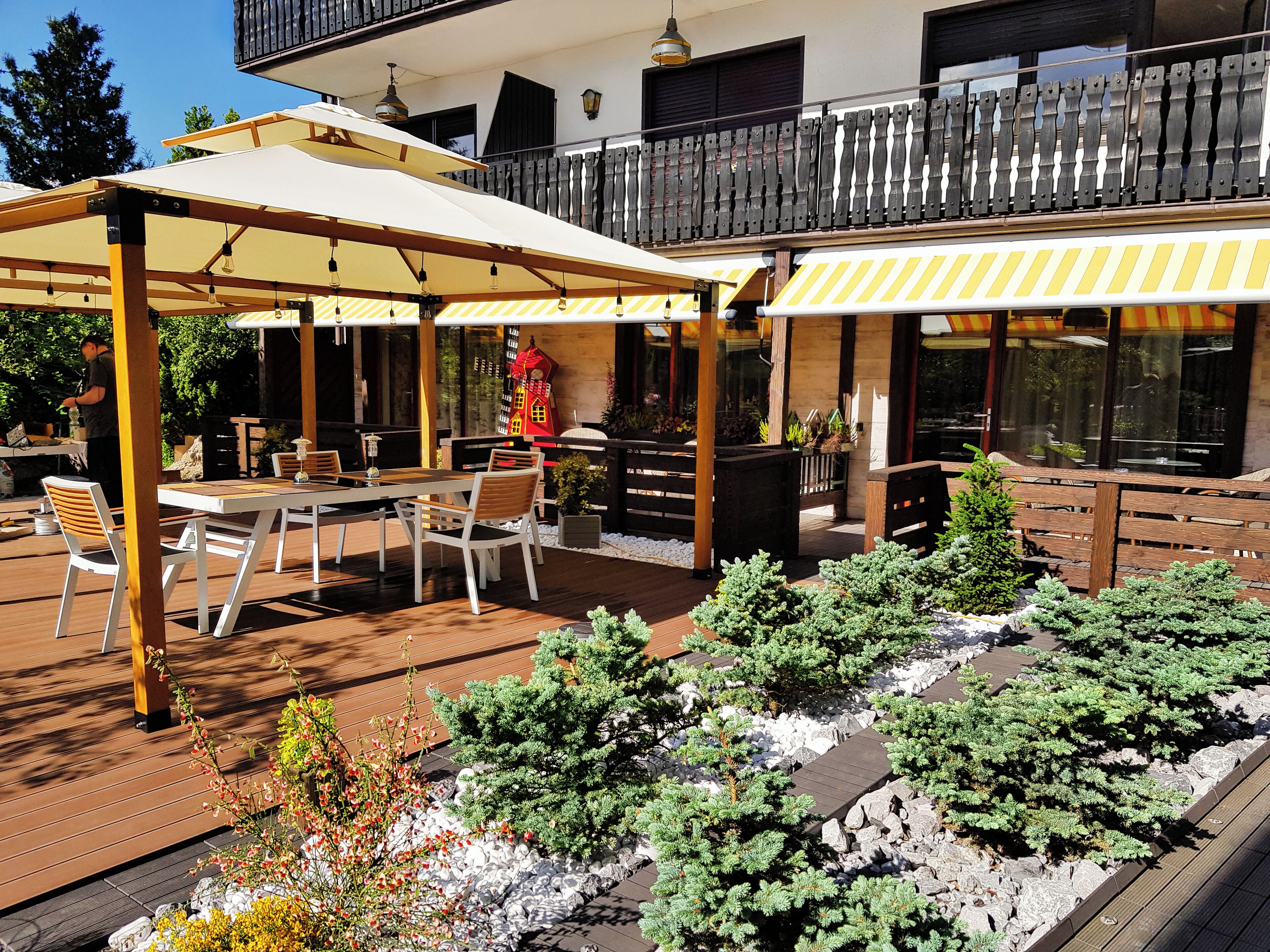 An outdoor patio with wooden tables and chairs, a covered seating area, and a decorative garden with plants and rocks.