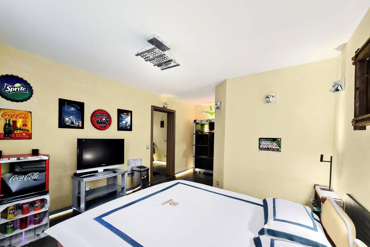Habitación de hotel con cama, televisor, mini bar y detalles decorativos, incluyendo una lata de Sprite y un cartel de Harley-Davidson.