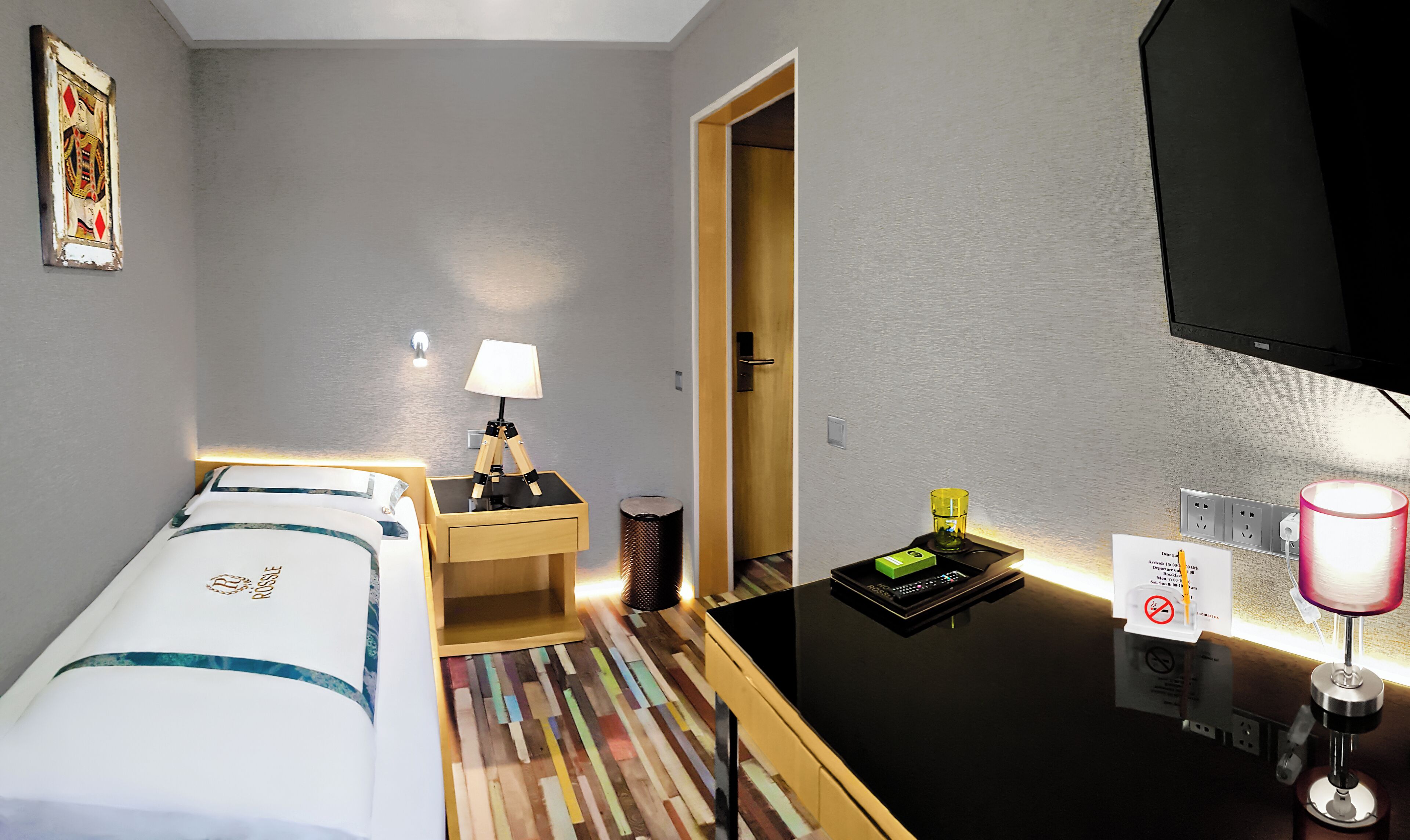 Una camera d'albergo con un letto, una scrivania, una lampada, un cestino, una TV e un tavolino con il telecomando.