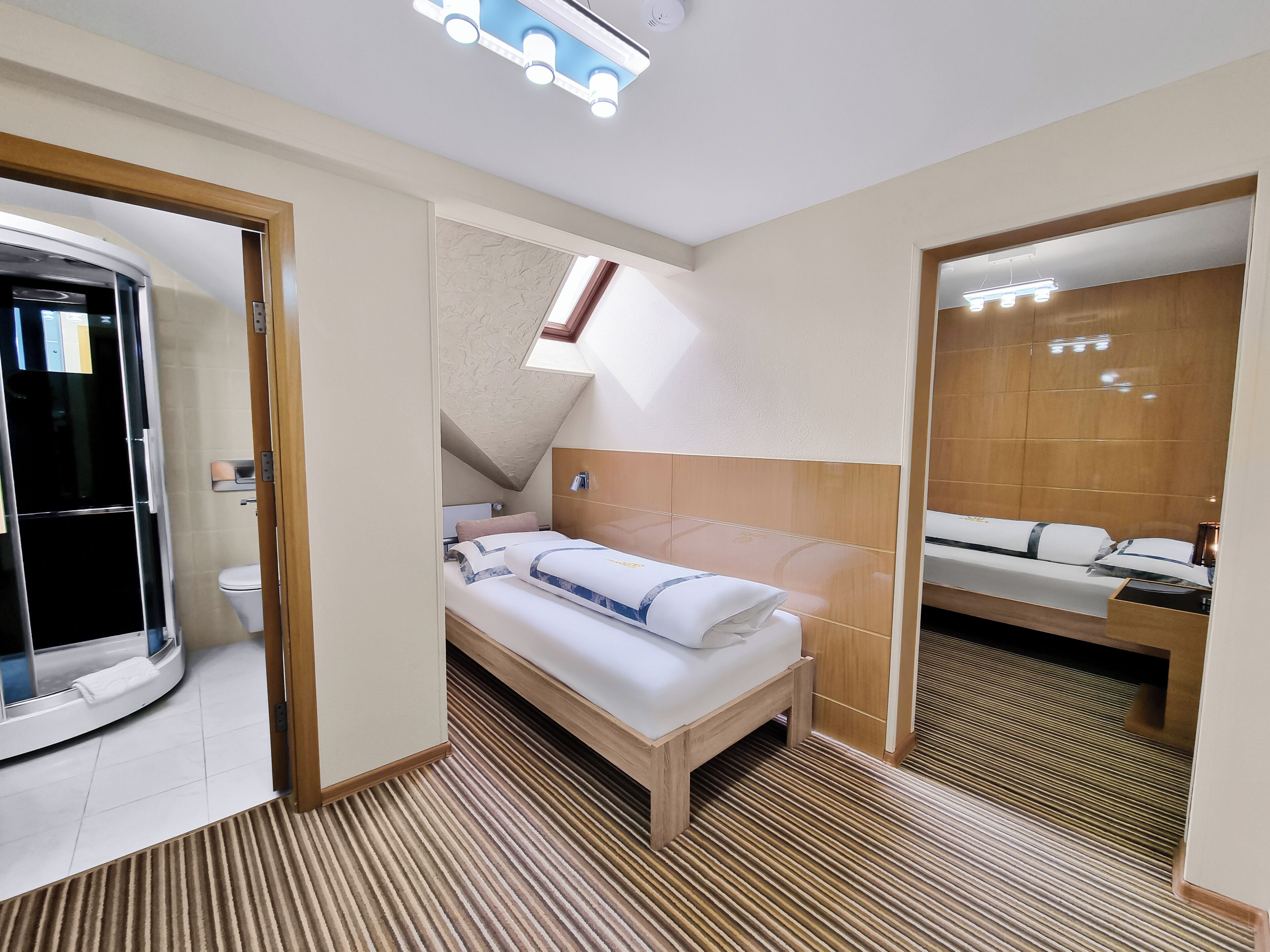A modern hotel room with a bed, a wooden headboard, a small skylight, and a bathroom with a toilet and shower.