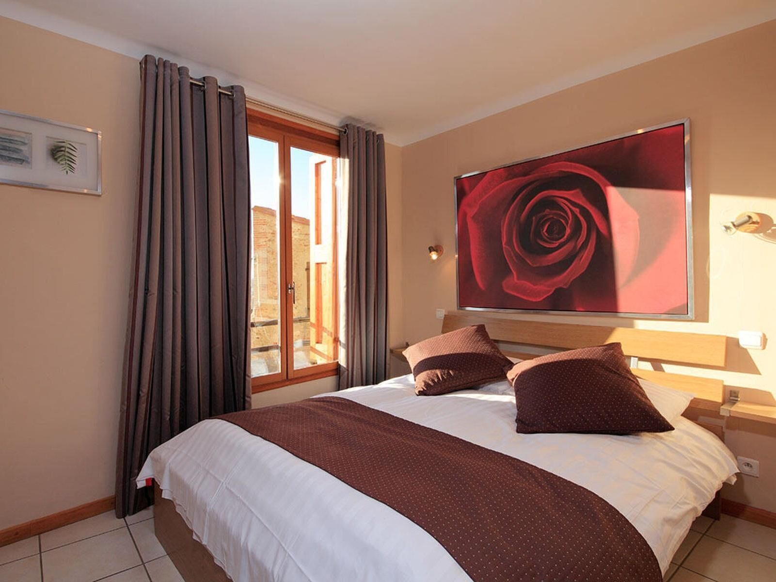 A hotel room with a large bed, a window with curtains, a framed picture on the wall, and a large painting of a rose.