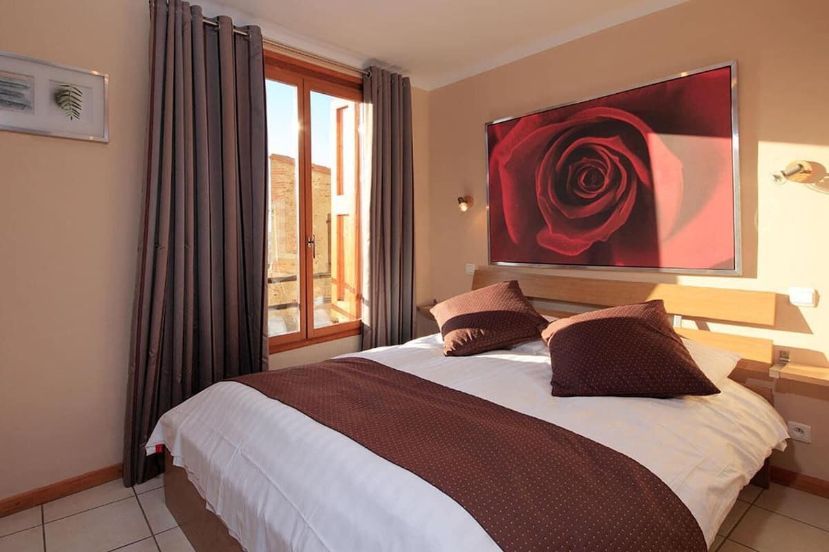 A hotel room with a large bed, a window with curtains, a framed picture on the wall, and a large painting of a rose.