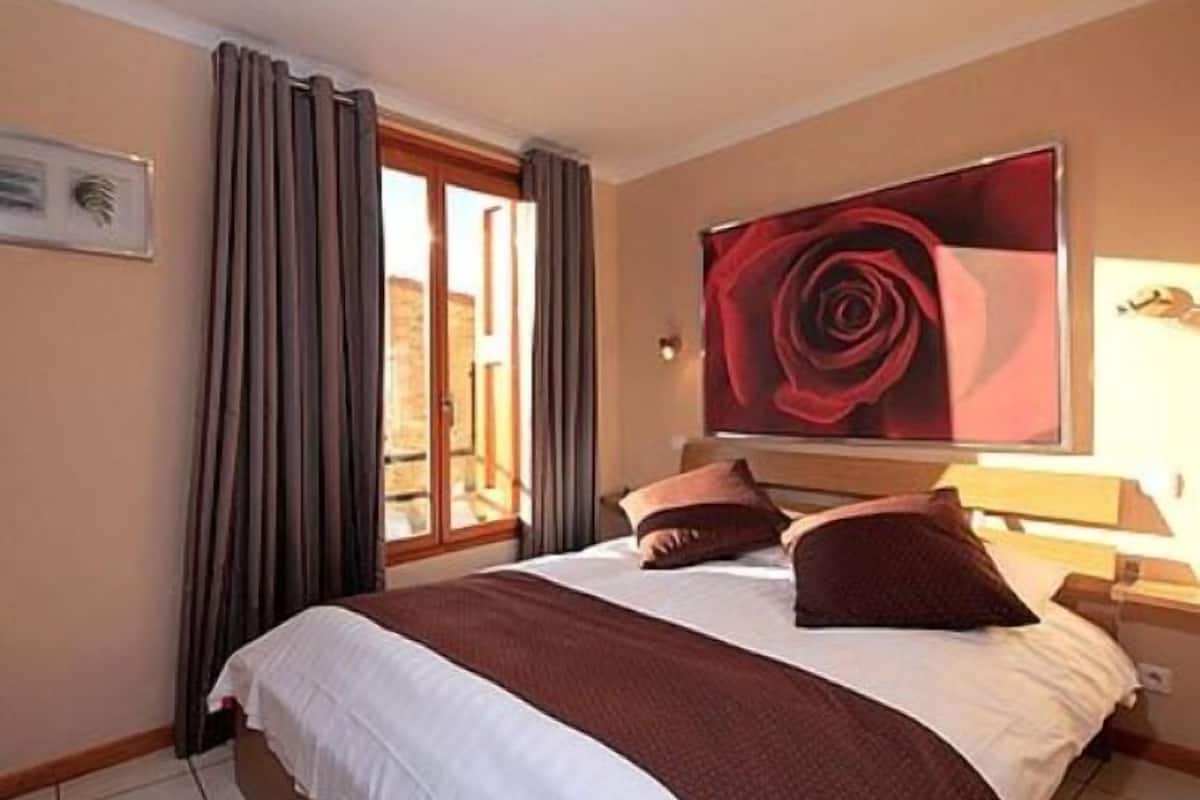 A hotel room with a large bed, a painting of a red rose on the wall, and a view of the outside through the window.
