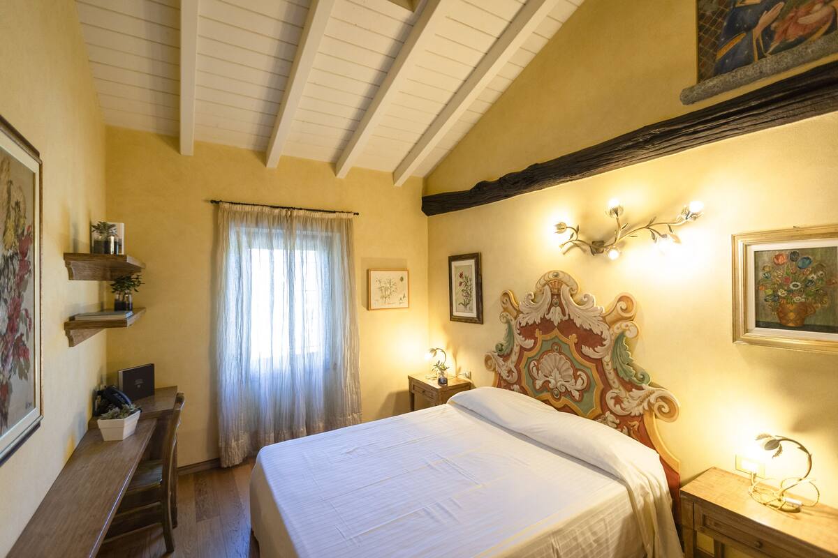 A bedroom with a bed, a desk, a chair, a nightstand, and a window with curtains.