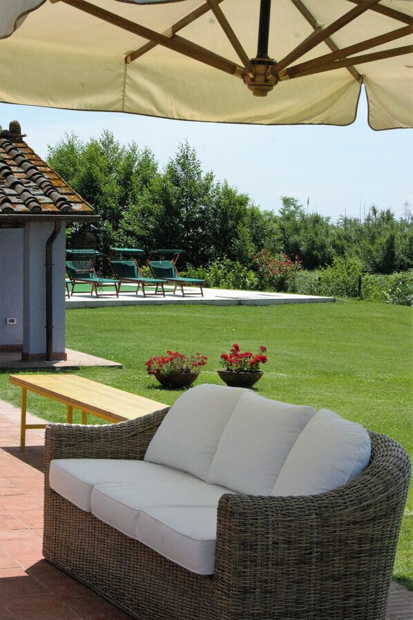 Un espacio al aire libre con un sofá de mimbre, una mesa de madera y un banco. Hay dos macetas con flores rojas.
