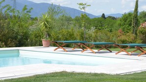 Uma piscina com espreguiçadeiras sob um toldo, rodeada por vegetação e montanhas ao fundo.