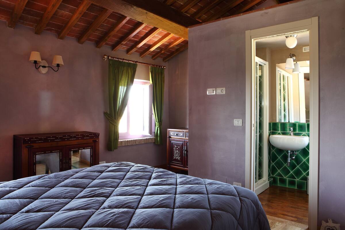 A bedroom with a bed, a wooden dresser, a window with green curtains, and a bathroom with a green tiled wall and a white sink.