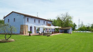Uma casa grande com um amplo gramado, área de estar externa com guarda-sóis e uma construção pequena.