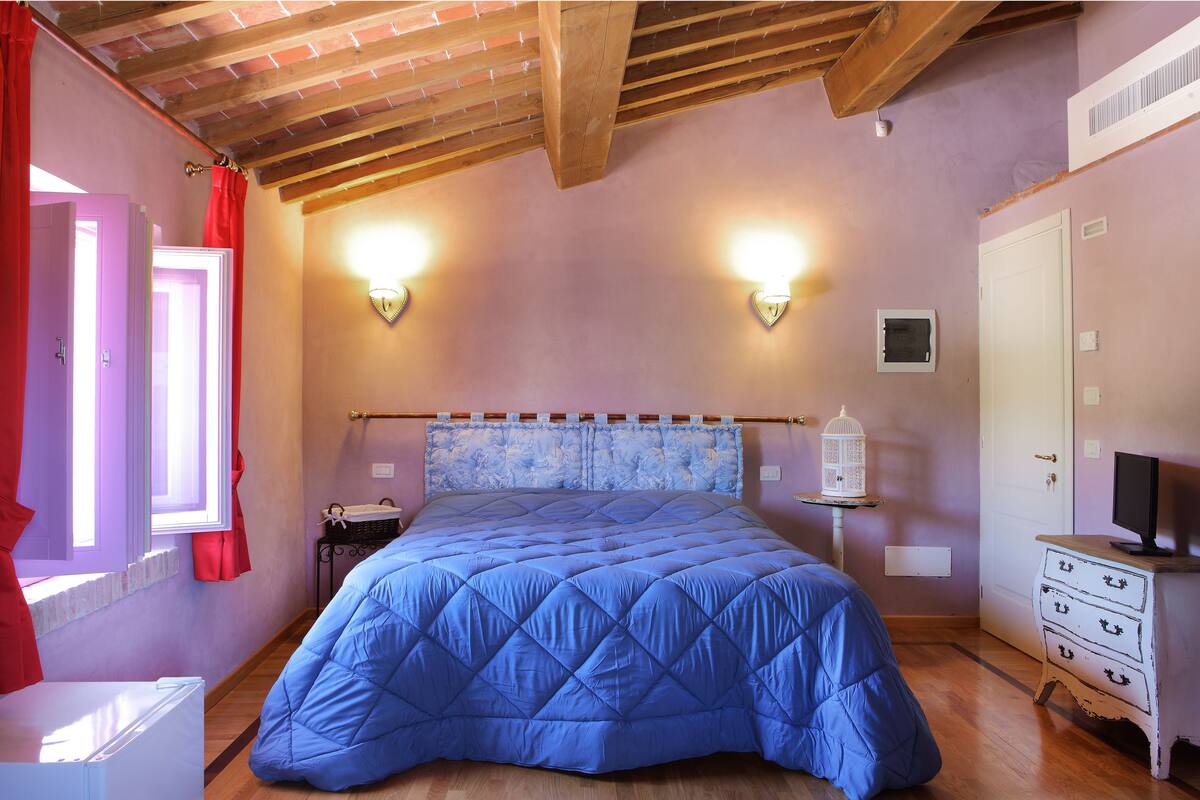 A bedroom with a blue quilted bedspread, a wooden ceiling, a white dresser, and a nightstand with a lamp.