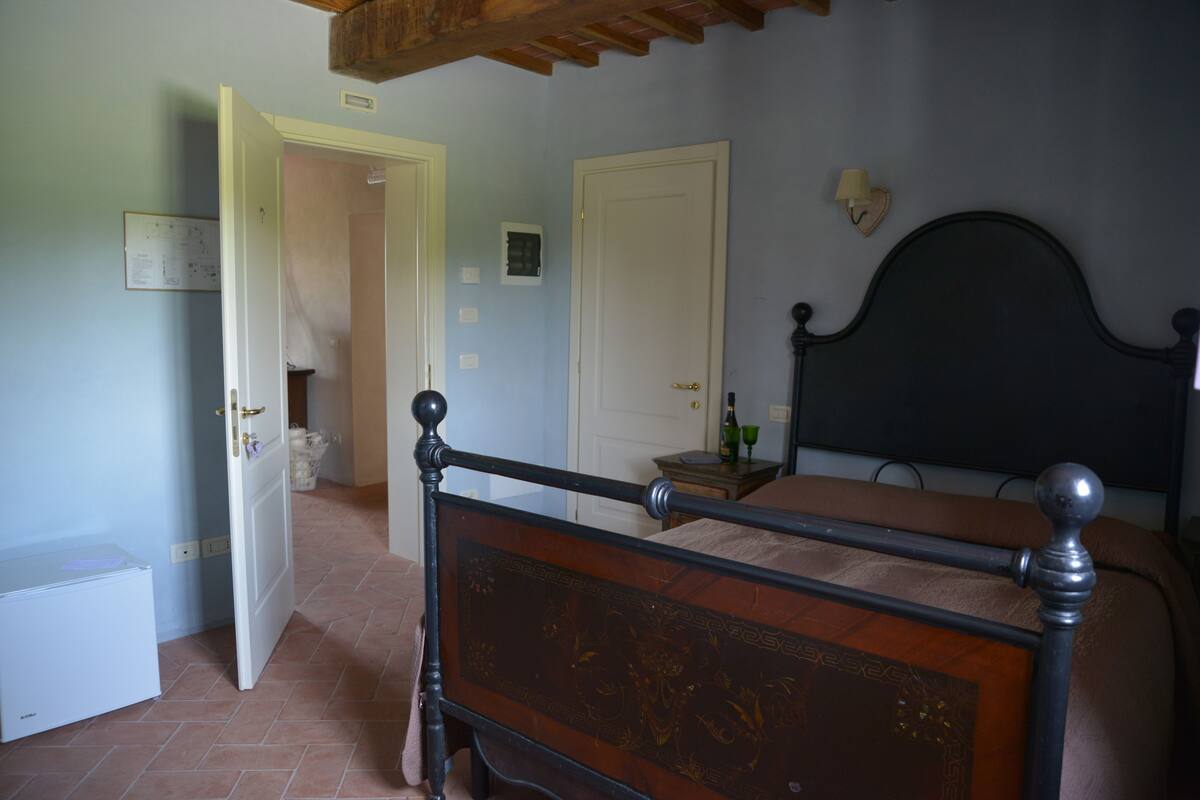 A bedroom with a wooden bed frame, a white door, a small bedside table with a lamp, and a tiled floor.