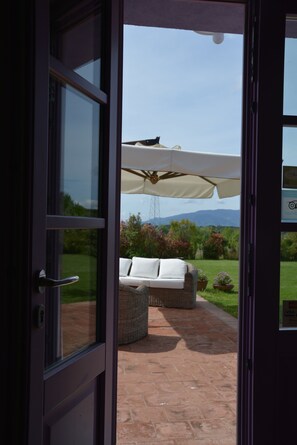 Puerta abierta que da a un patio exterior con un paraguas blanco, muebles de mimbre y vistas a la vegetación y montañas.
