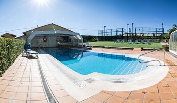 Una piscina con sombrilla, sillas de descanso y una cancha de tenis al fondo.