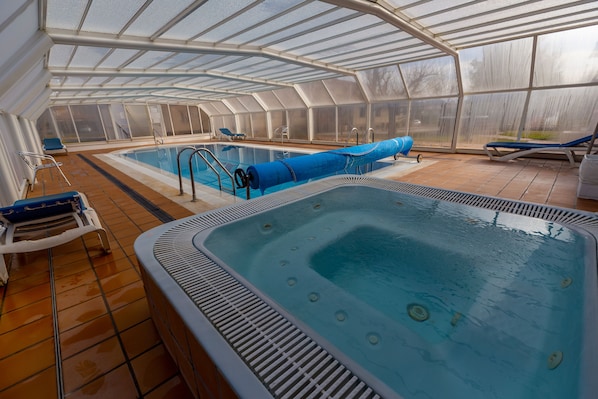 A covered indoor swimming pool with a hot tub, lounge chairs, and a sunshade structure.