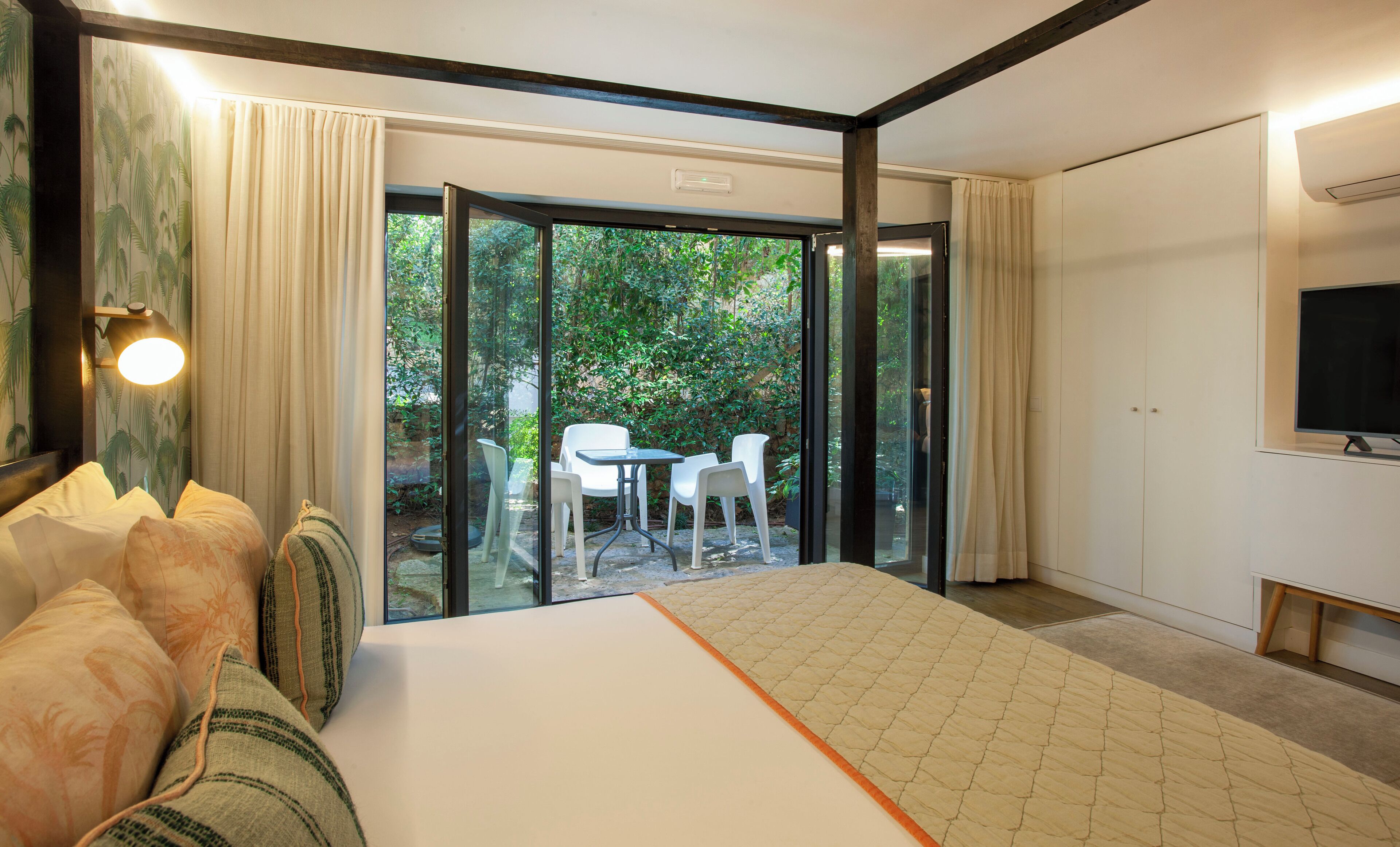 Habitación de hotel con cama, sofá, televisión y puerta corrediza de vidrio que da a un patio con mesa y sillas.