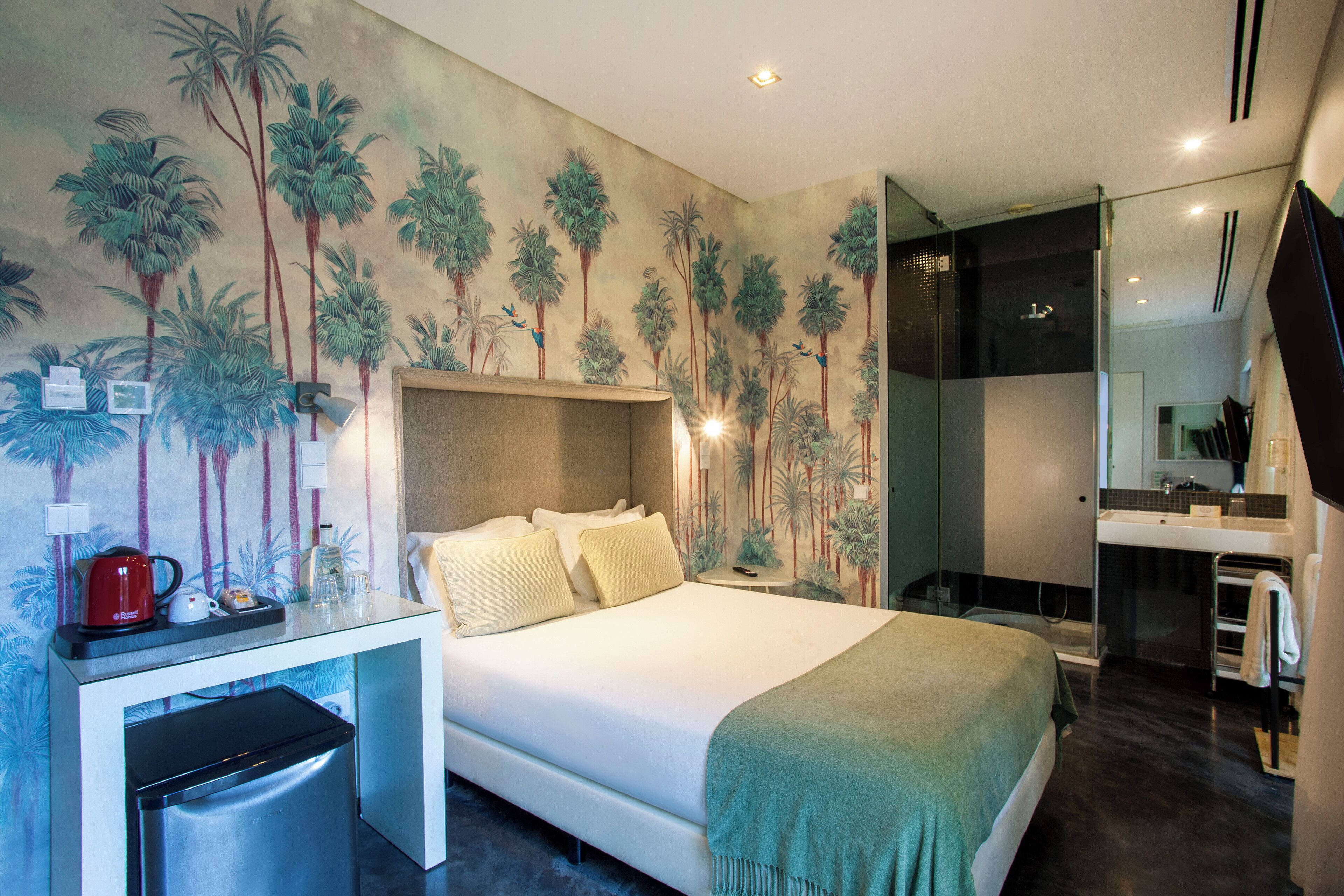 Een moderne hotelkamer met een bed, een bureau met koffiezetapparaat, een spiegel en een badkamer met wastafel en spiegel.