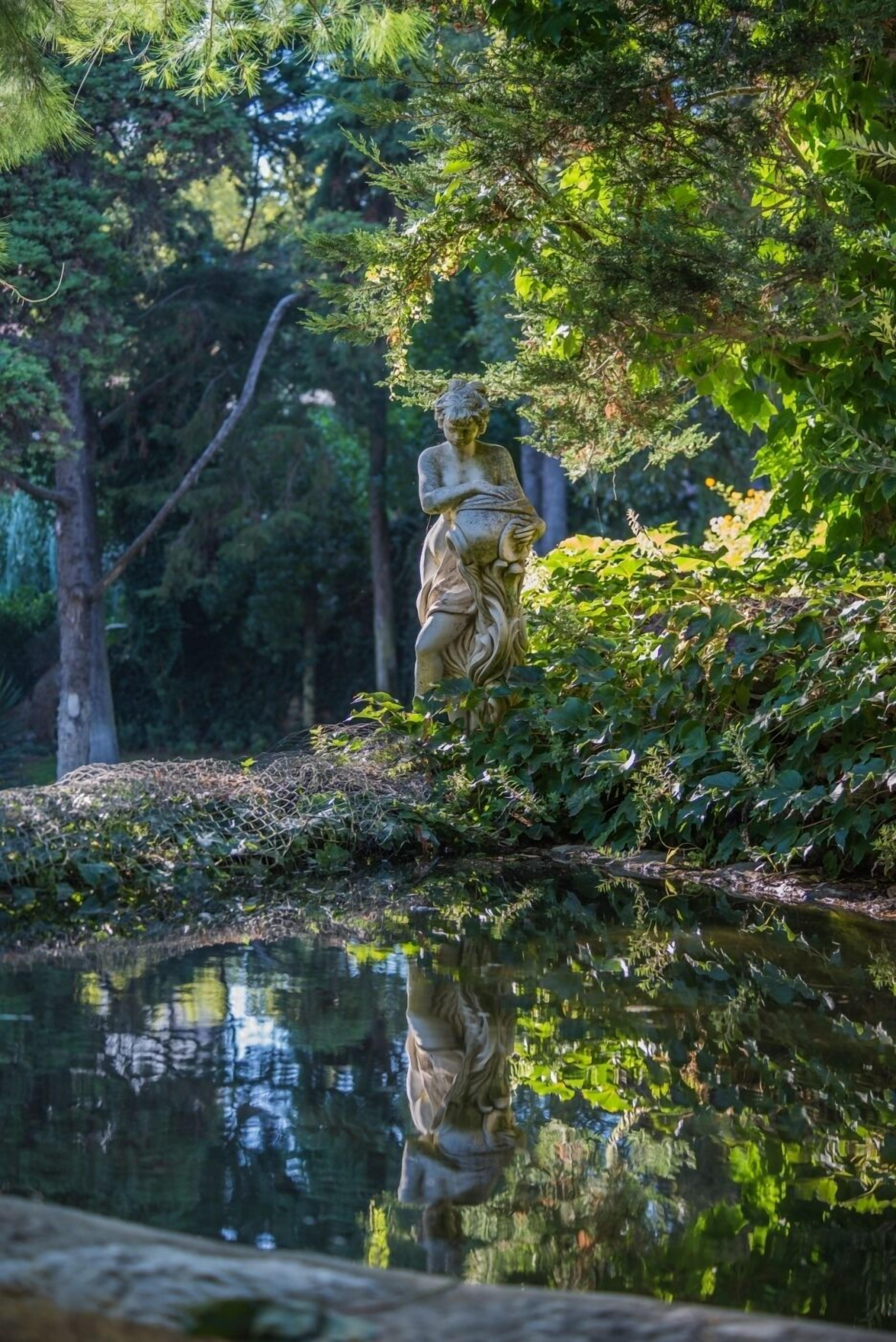 A statue of a lioness on a stone pedestal by a pond, surrounded by lush greenery.