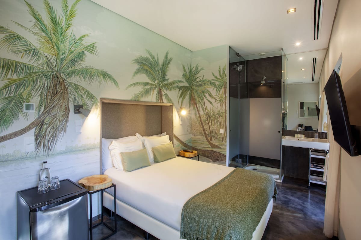 Una habitación de hotel moderna con una cama grande, una mesita de noche, una pequeña mesa de madera, cabina de ducha con mampara de vidrio y un baño con espejo y lavabo.