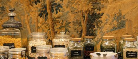 Une variété de bocaux en verre, étiquetés et contenant différents types d’aliments, comme des céréales, du chocolat et des graines, sont disposés sur une table devant un fond à motifs.