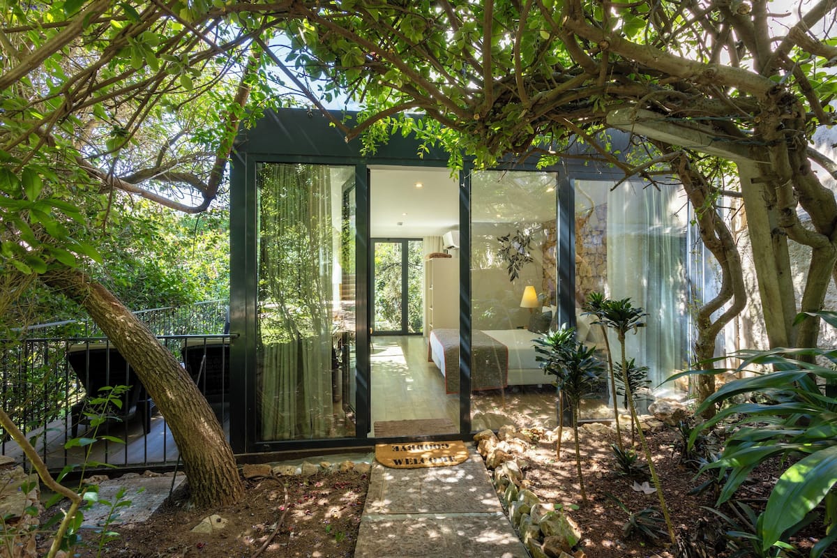 Una casa moderna de vidrio con cama, lámpara y felpudo en un jardín exuberante.