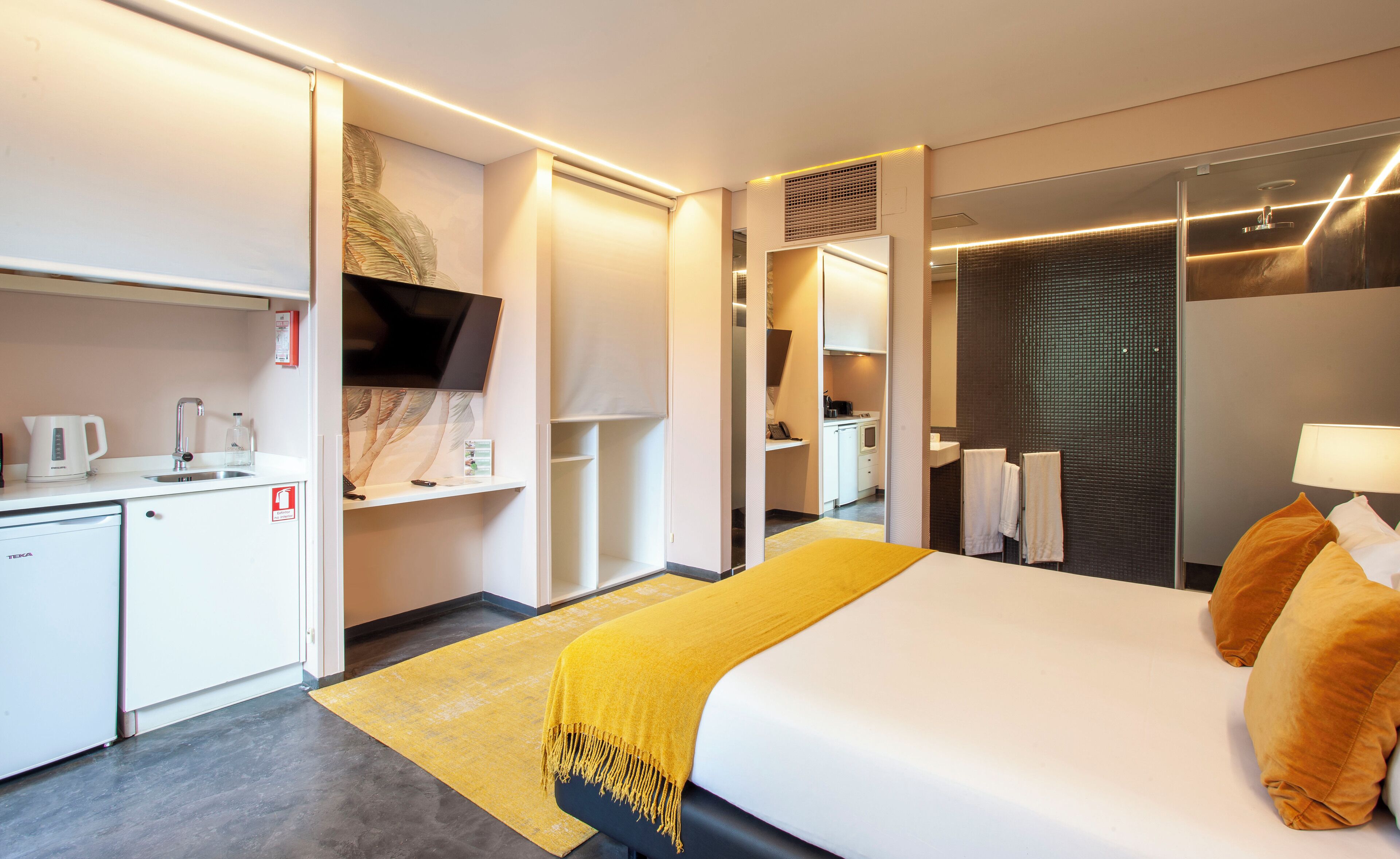 A modern hotel room with a bed, a desk with a kettle, a TV, and a bathroom with a shower.