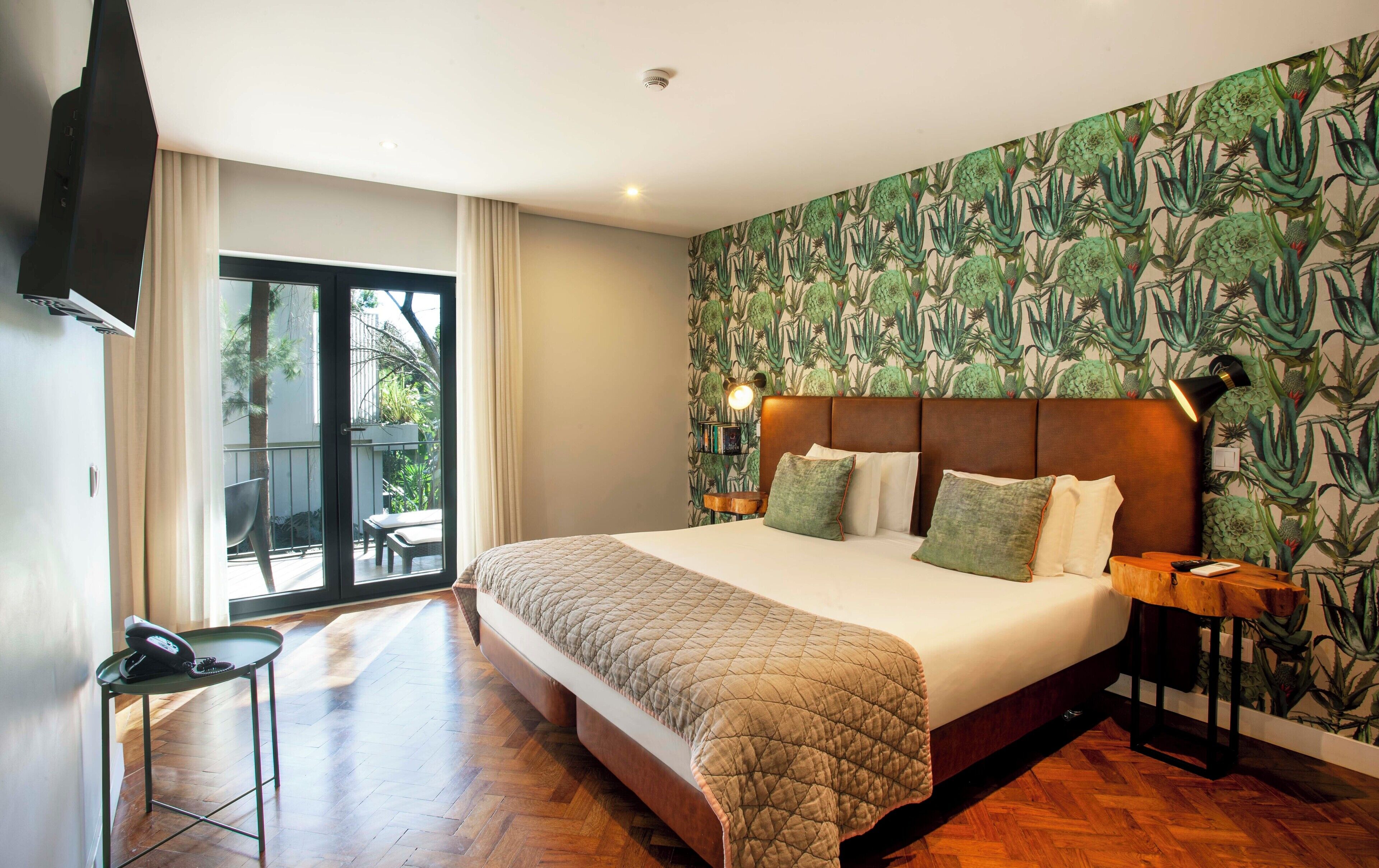 A bedroom with a large bed, a bedside table with a lamp, a small table with a telephone, and a sliding glass door leading to an outdoor area.
