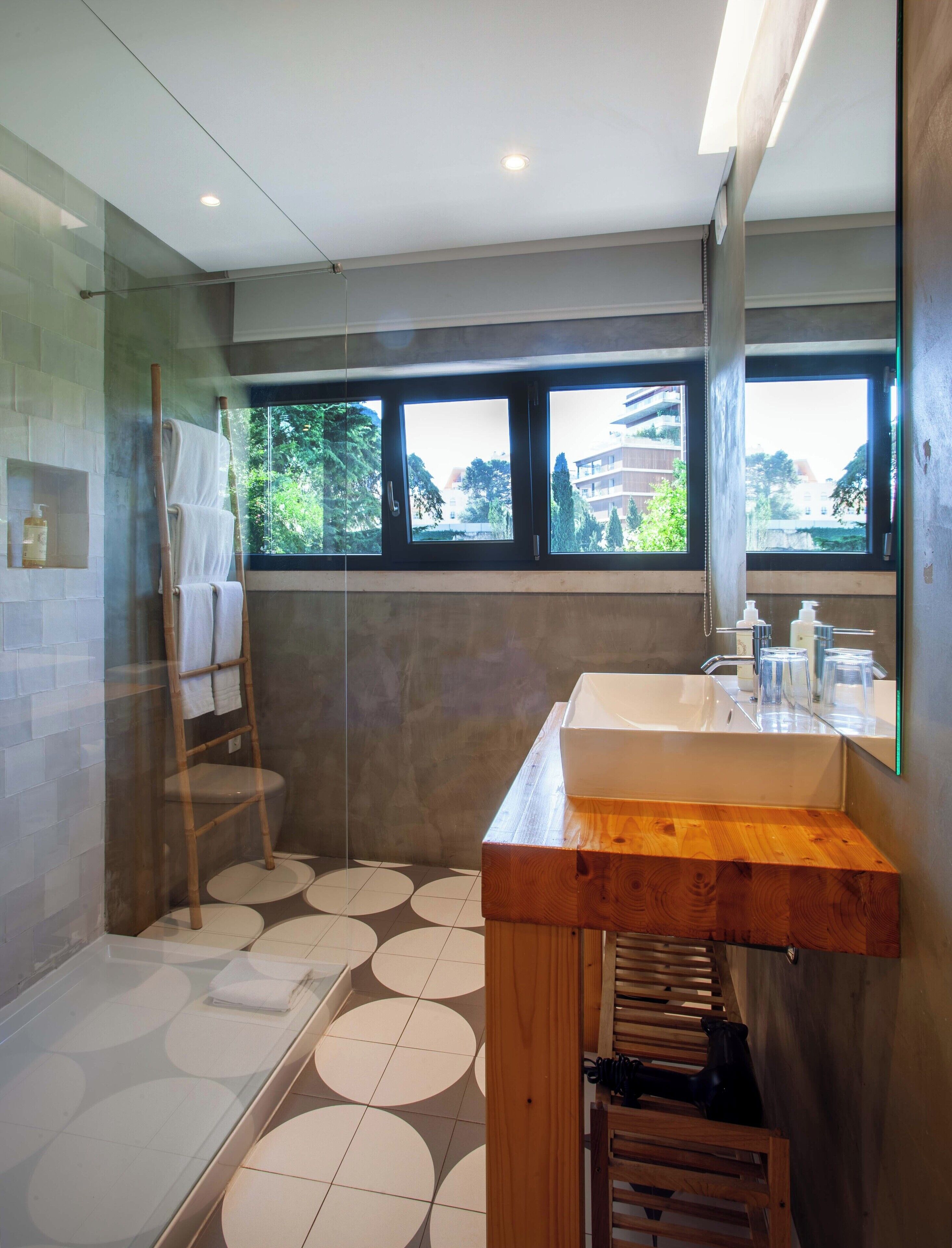 A modern bathroom with a wooden vanity, a glass shower enclosure, and a view of buildings through the windows.