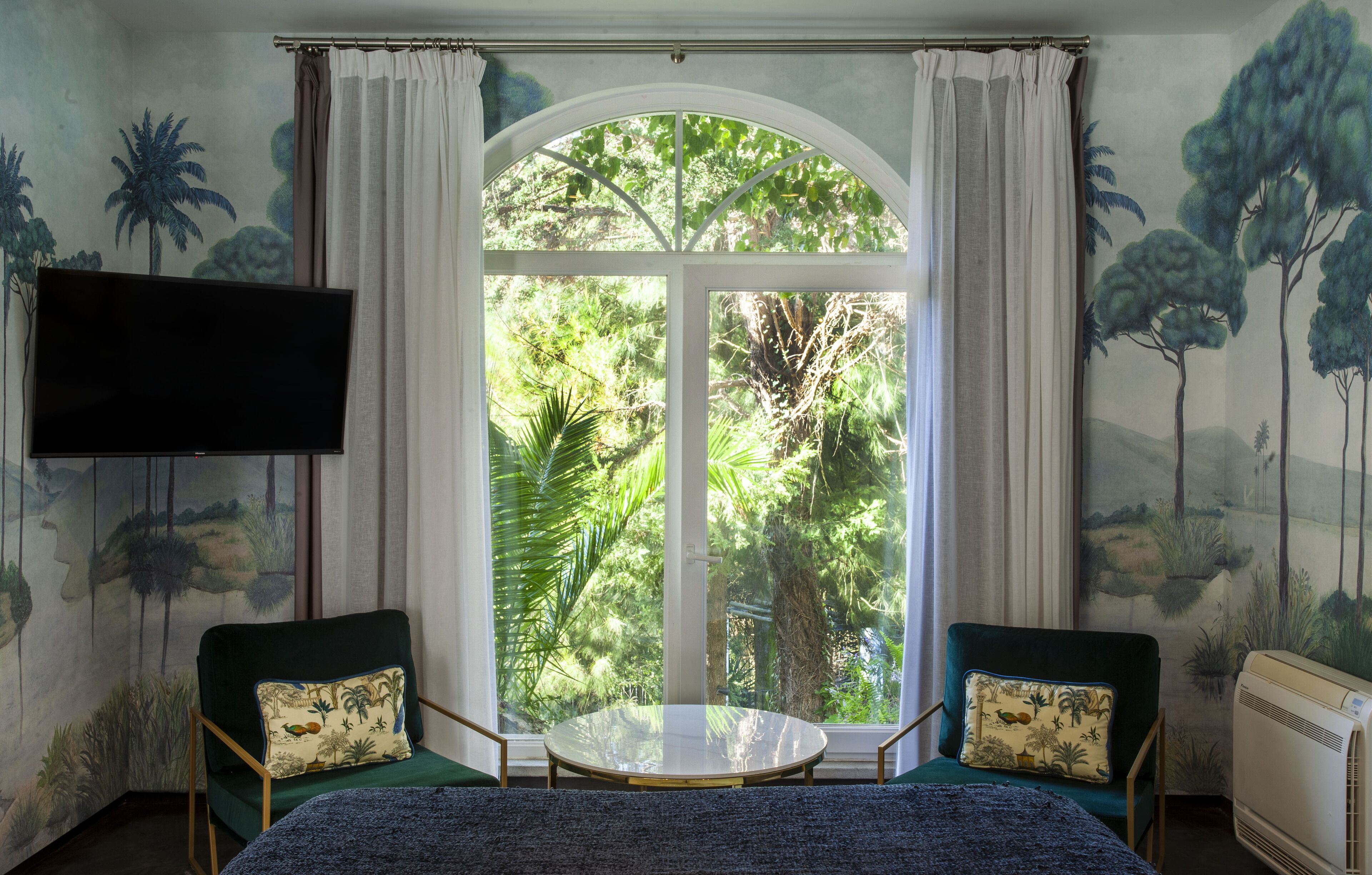 A room with a large arched window, two armchairs with matching cushions, a flat-screen TV mounted on the wall, and a round table in the center.