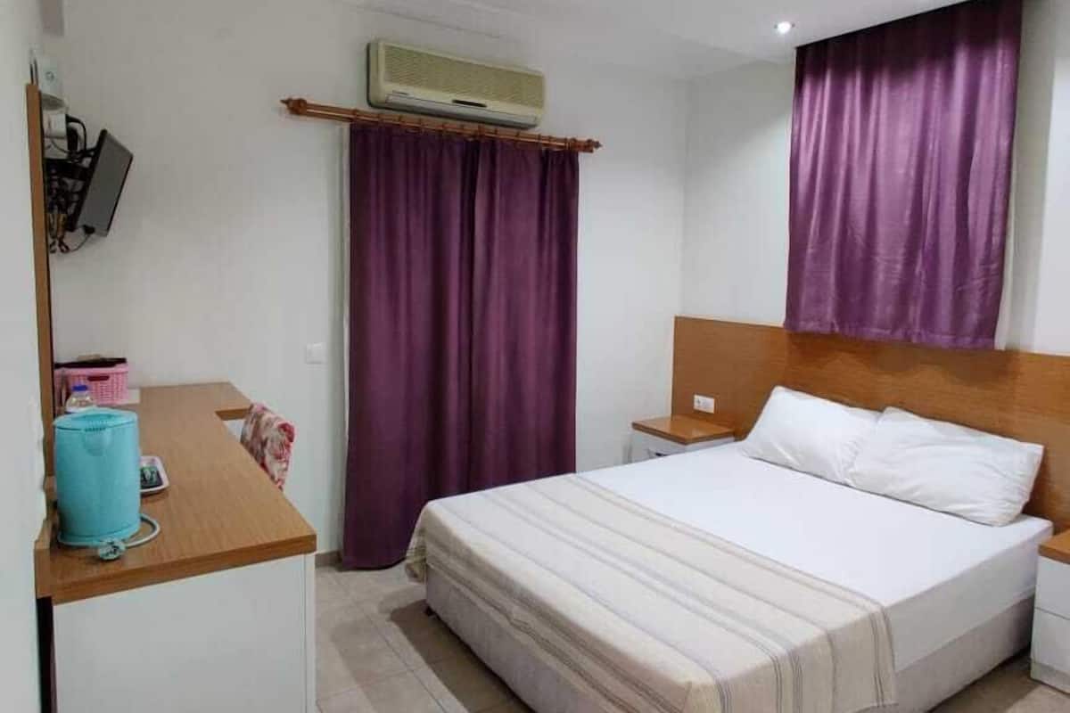 A hotel room with a bed, a desk, a chair, an air conditioning unit, and a window with purple curtains.