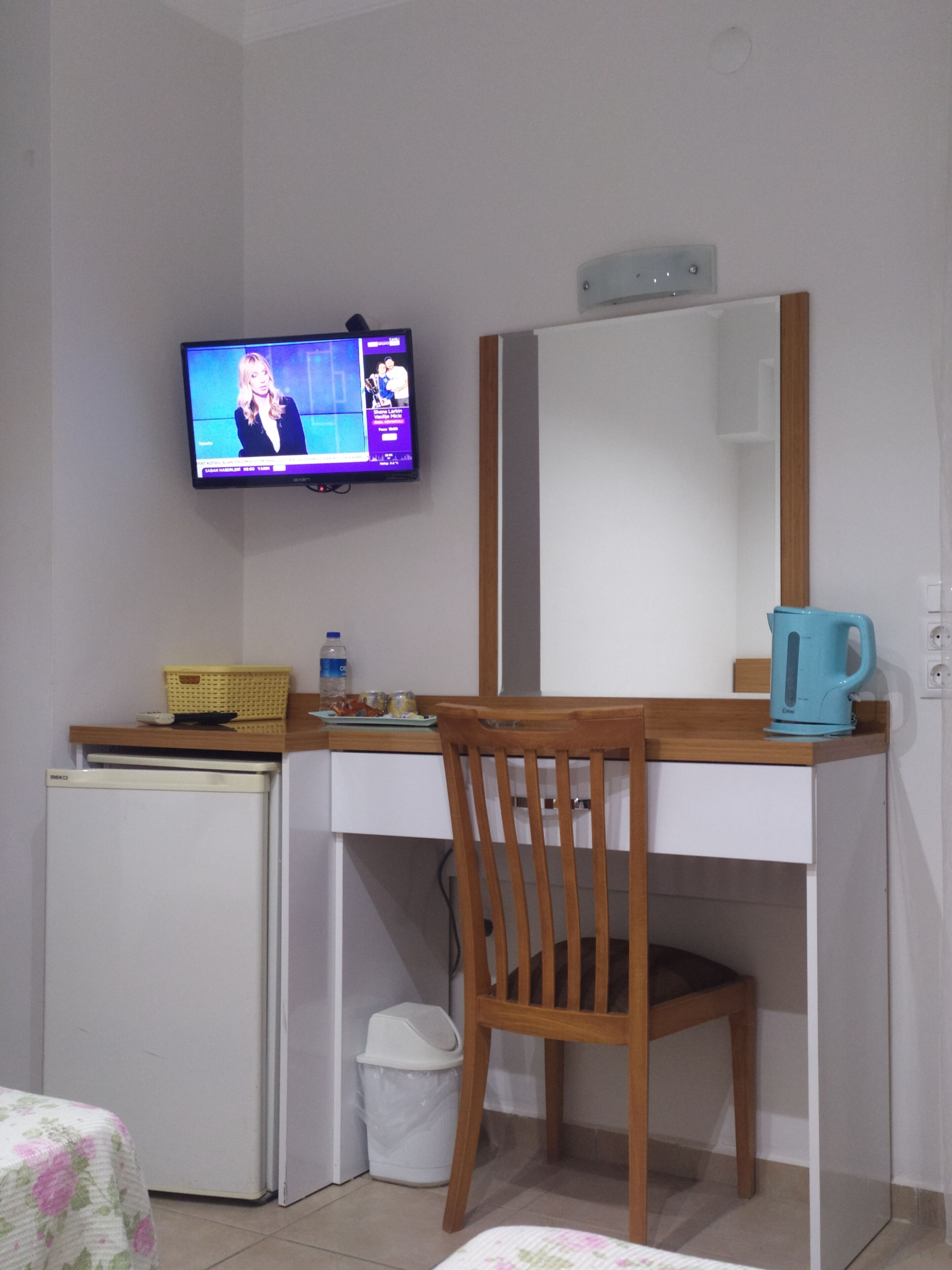 A compact room with a wooden desk, a television mounted on the wall, a small refrigerator, a trash bin, and a bed with a floral bedspread.