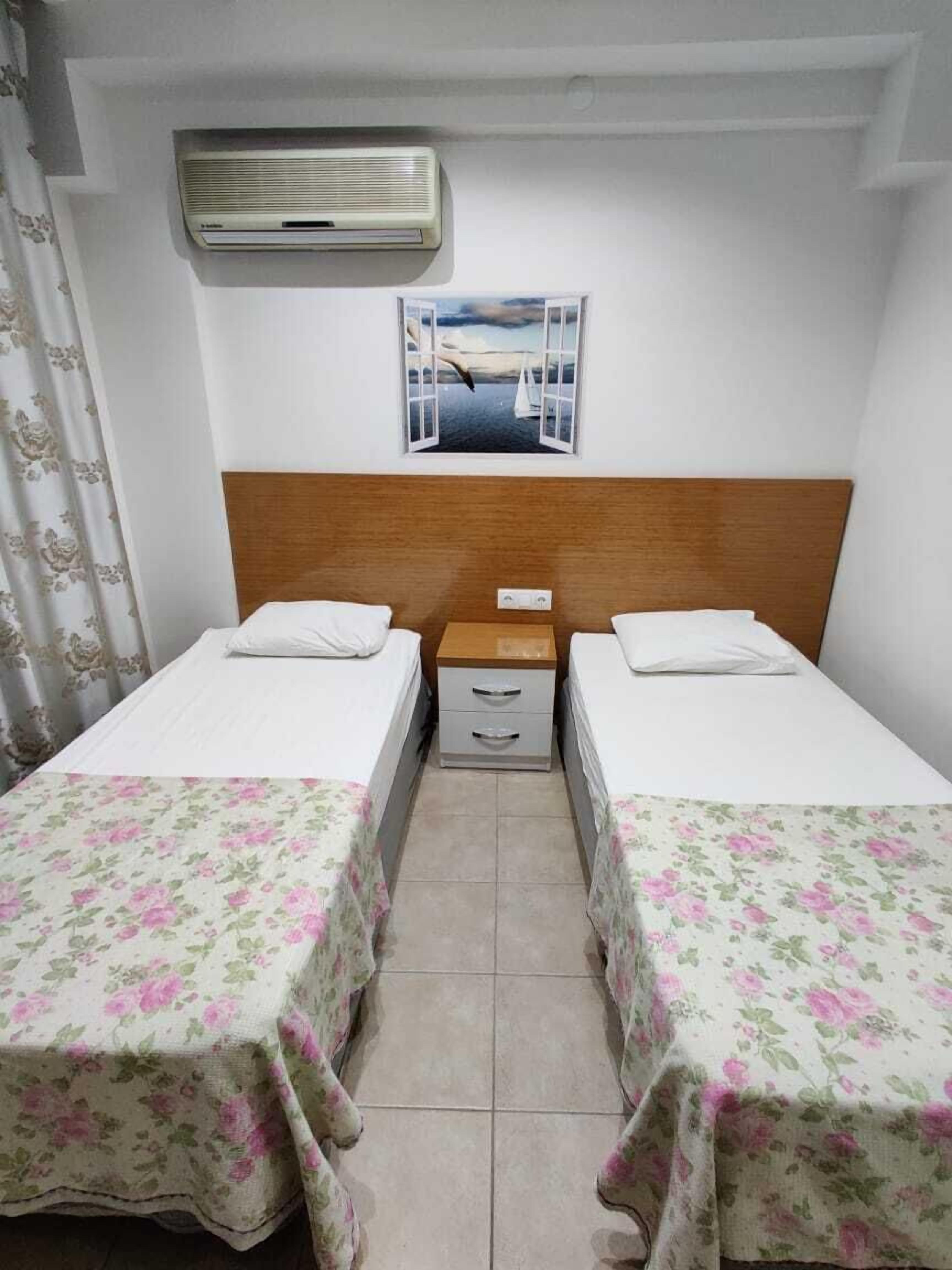 Une petite chambre propre, avec deux lits simples, une tête de lit en bois, une petite table de chevet avec des tiroirs, une fenêtre donnant sur la mer et un climatiseur fixé au mur.