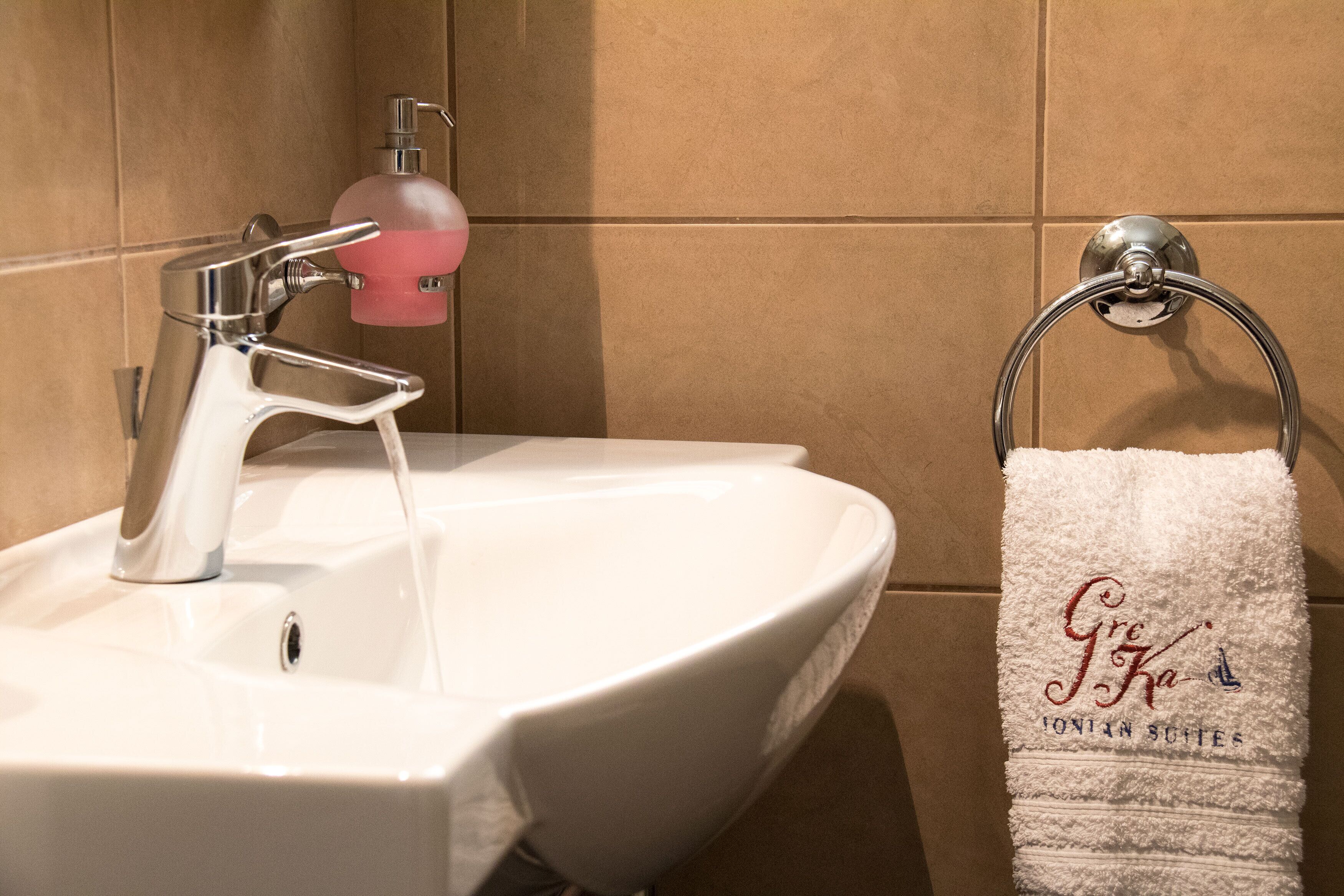 A bathroom sink with a modern faucet and a soap dispenser. A towel rack with a white towel displaying "Gre Ka" and "Ionian Suites" is visible.