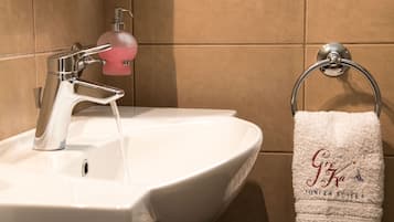 A bathroom sink with a modern faucet and a soap dispenser. A towel rack with a white towel displaying "Gre Ka" and "Ionian Suites" is visible.