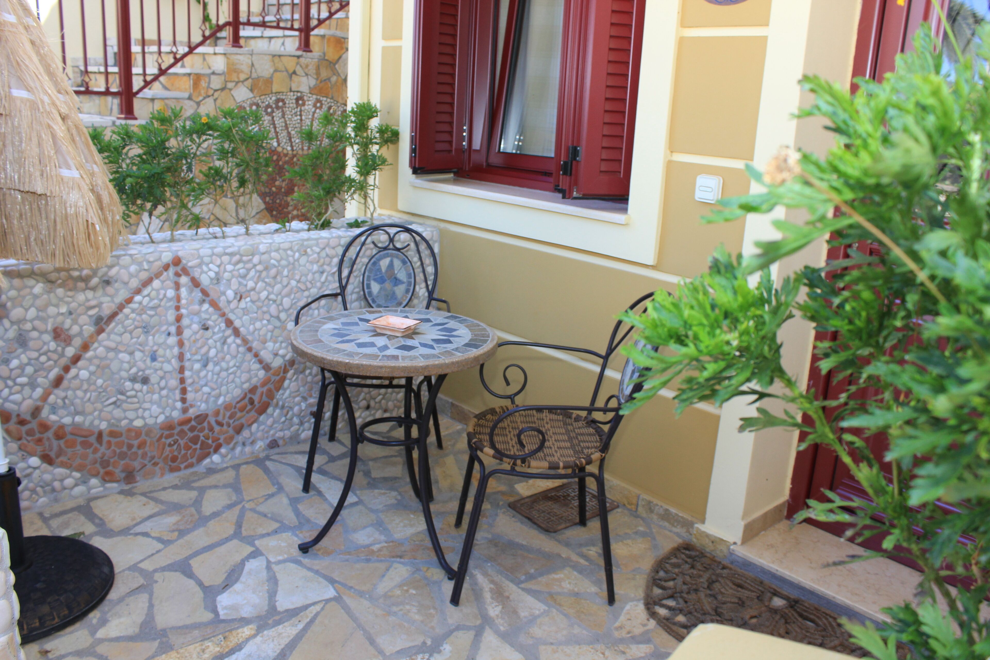 An outdoor patio with a mosaic-tiled floor, a round table, two wicker chairs, a thatched umbrella, and a window with red shutters.