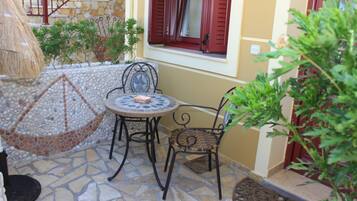 An outdoor patio with a mosaic-tiled floor, a round table, two wicker chairs, a thatched umbrella, and a window with red shutters.