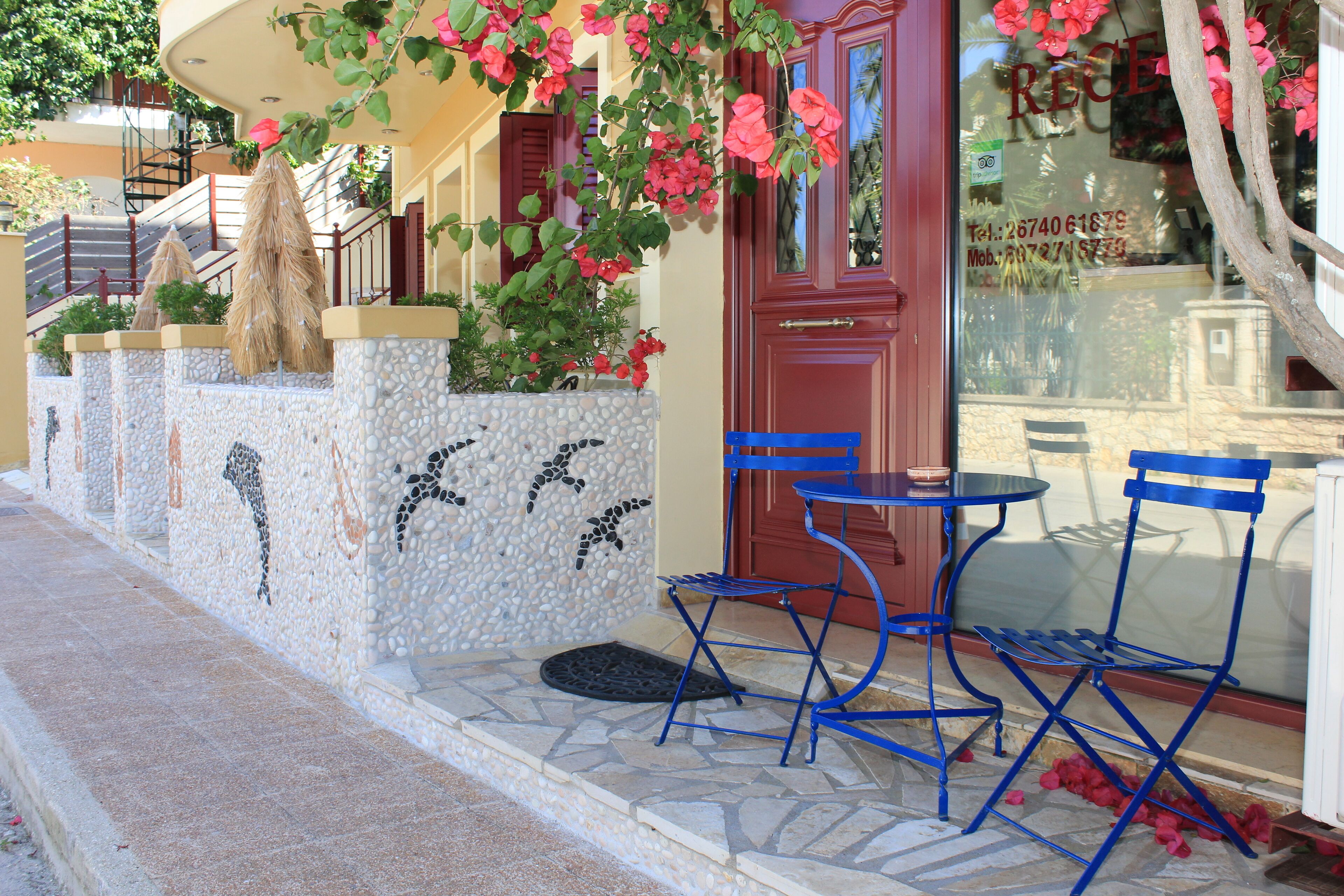 A hotel entrance with a stone wall, a red door, and blue chairs outside.