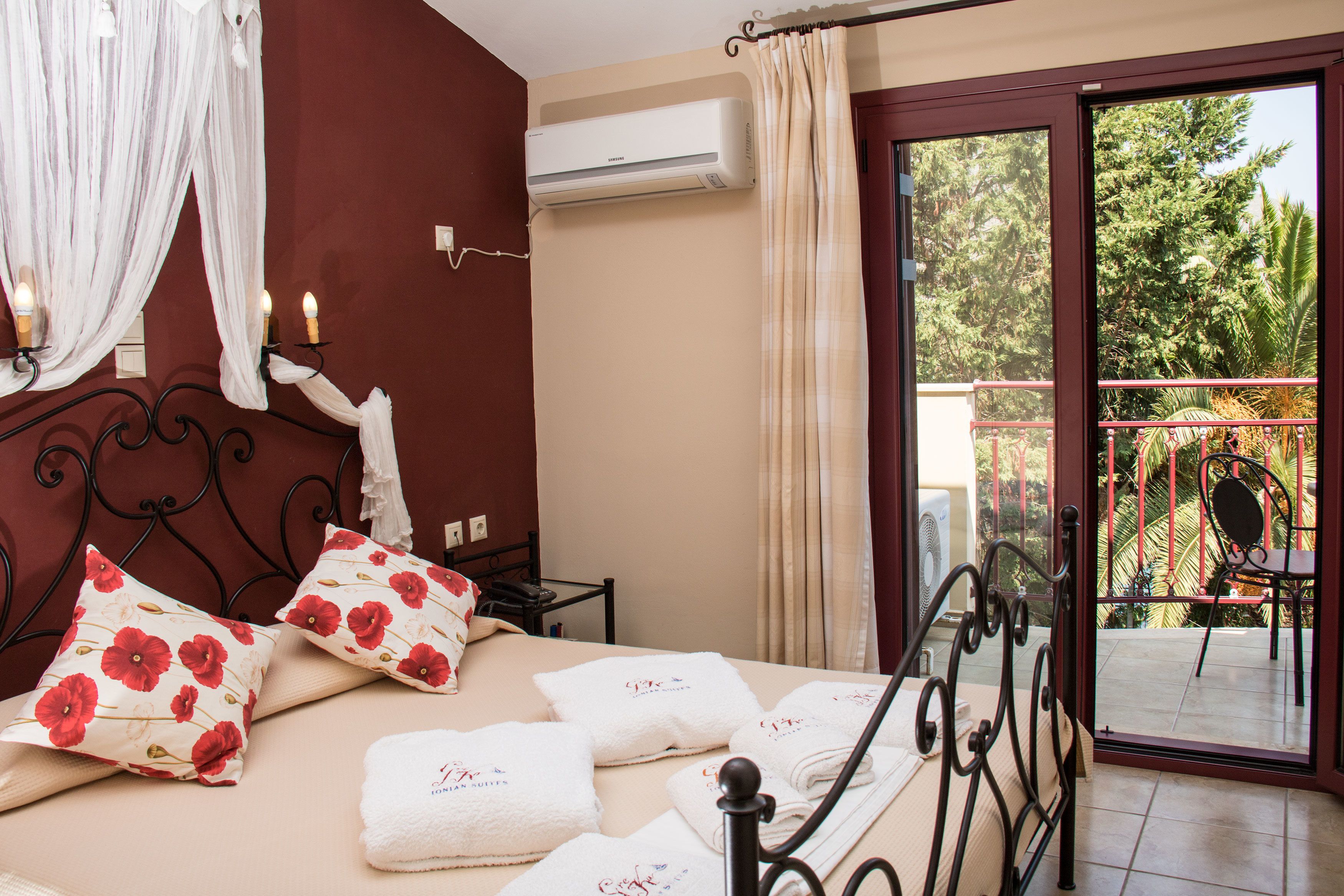 A hotel room with a bed, pillows, a bedside table, an air conditioning unit, and a view of a balcony with chairs and plants.
