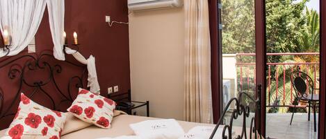 A hotel room with a bed, pillows, a bedside table, an air conditioning unit, and a view of a balcony with chairs and plants.