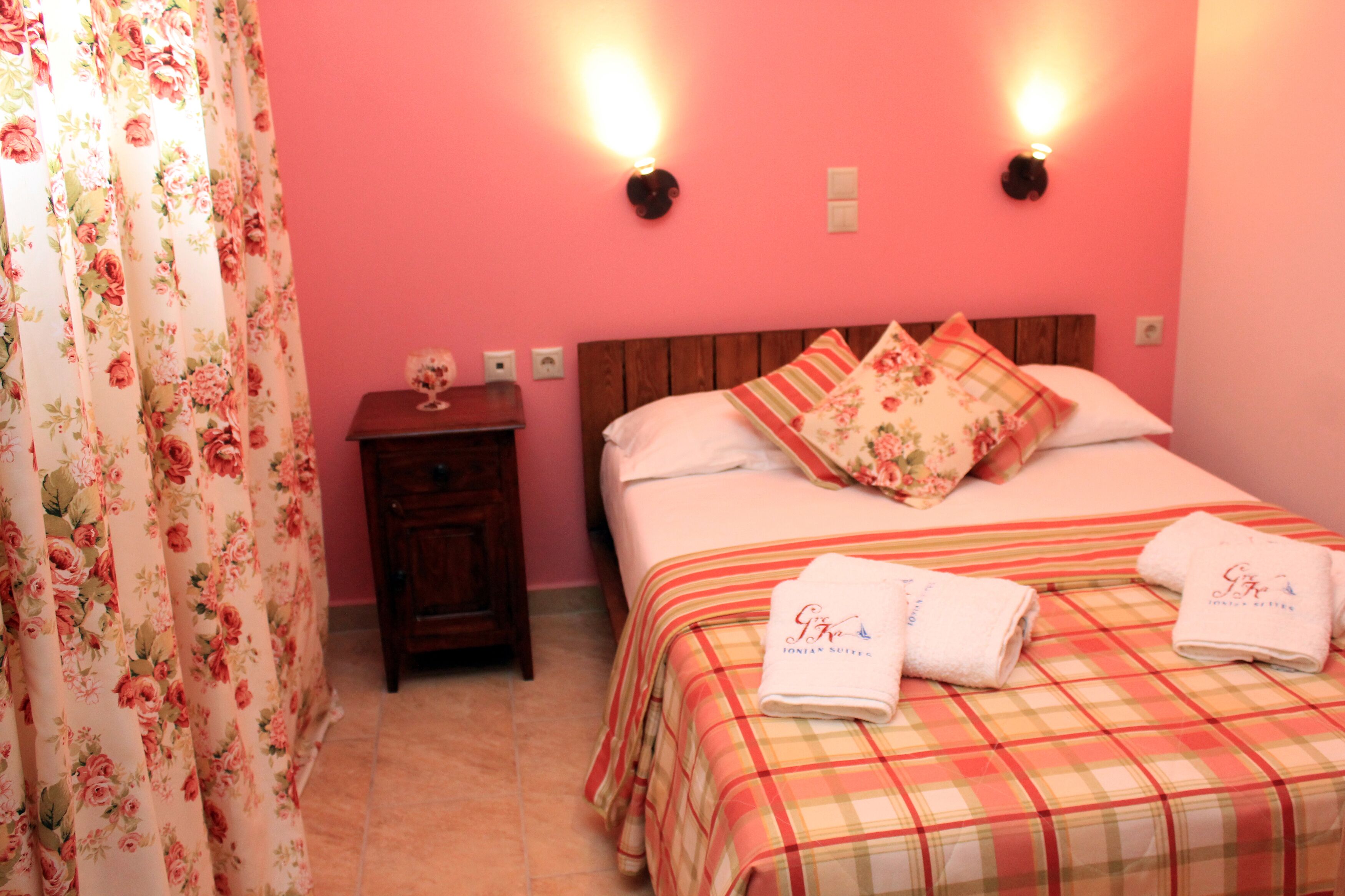 Un dormitorio con pared rosa, cortinas florales, una mesita de noche de madera, cama con ropa de cama de cuadros y dos almohadas, y dos luces fijadas en la pared.