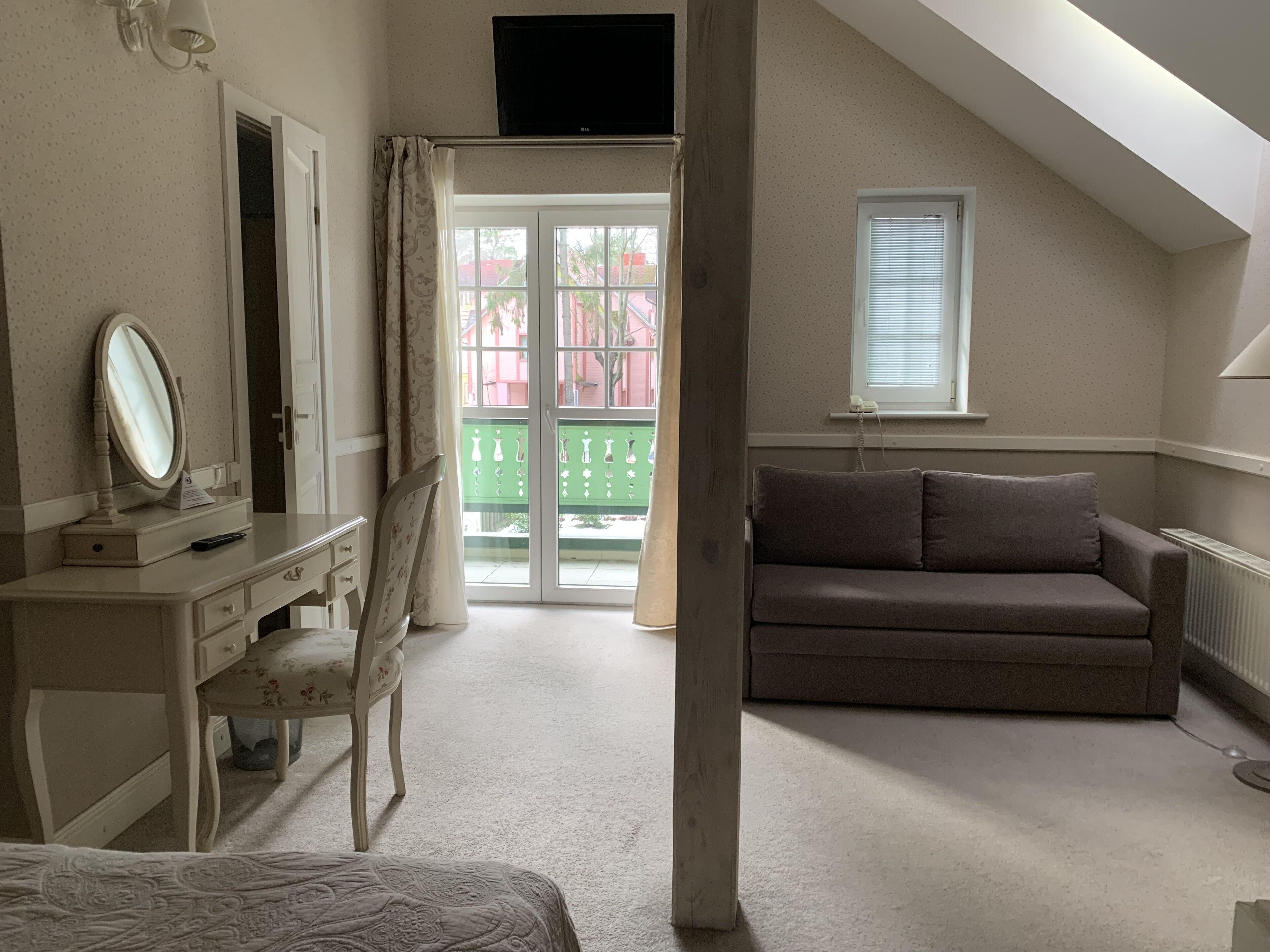A bedroom with a bed, a desk with a mirror, a dresser, a brown sofa, a window, and a balcony with a green railing.