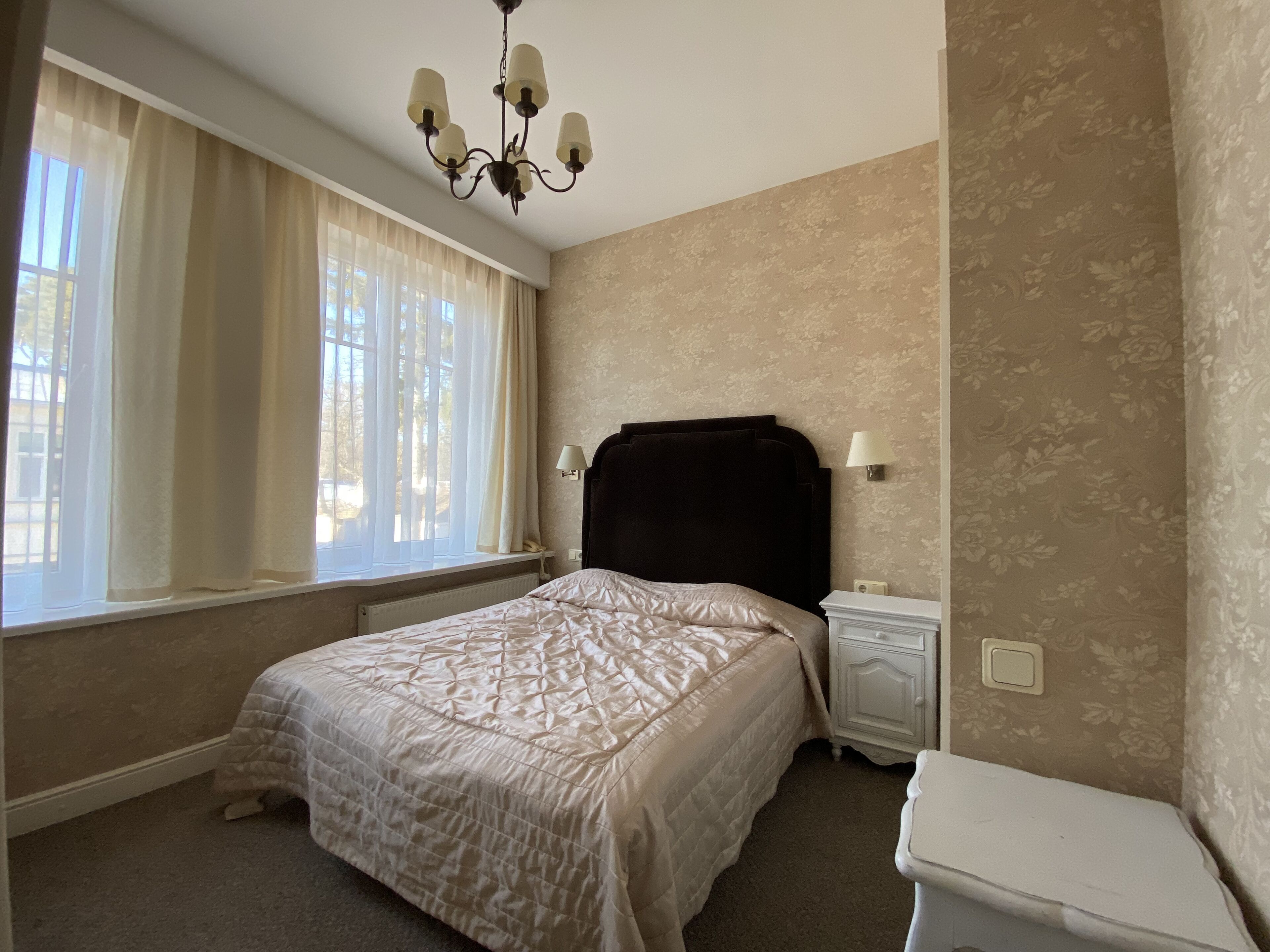 Un dormitorio con cama, mesitas de noche, lámpara, candelabro y ventana con cortinas.