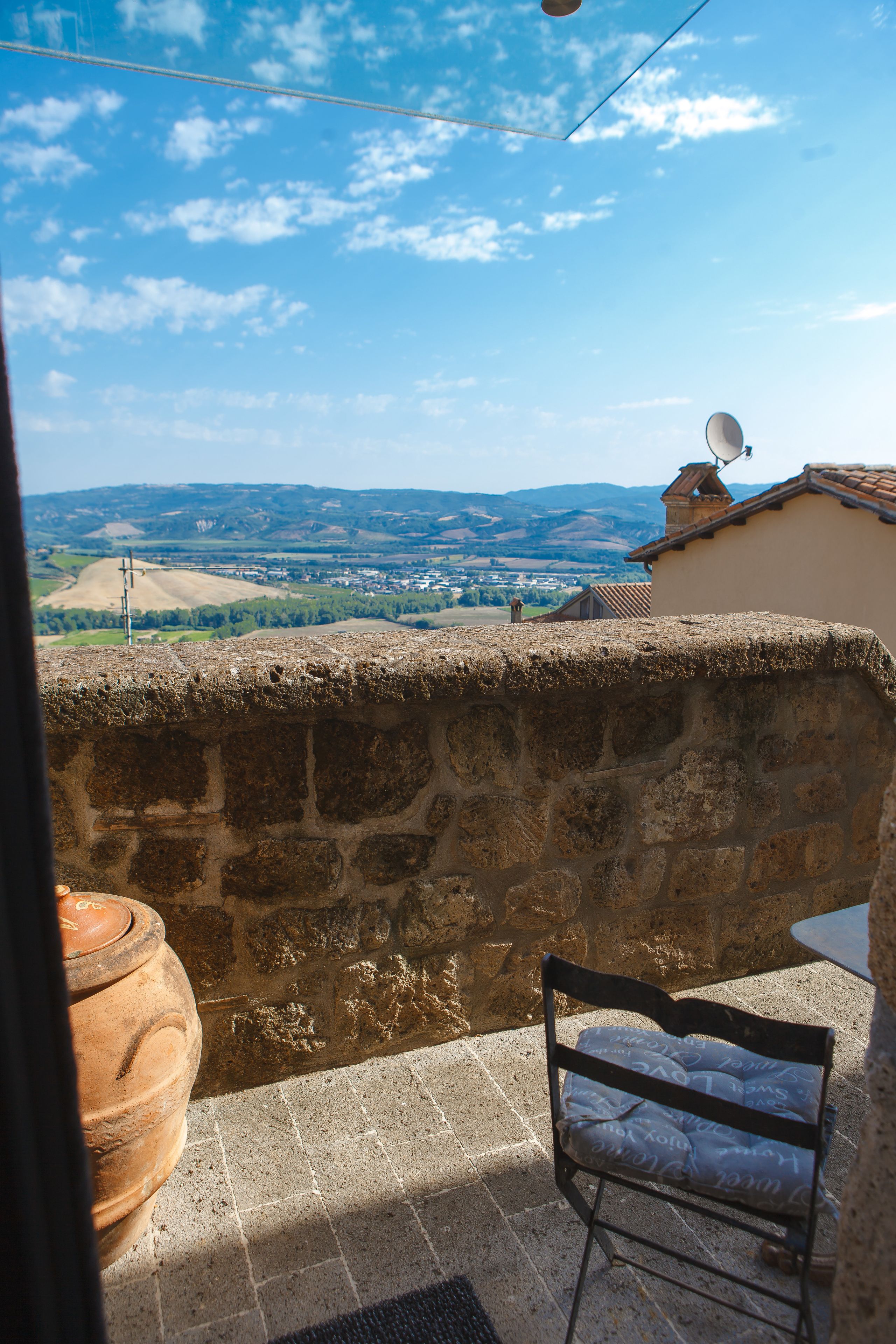 A stone wall with a view of a town, a rustic bench, and a large clay pot.