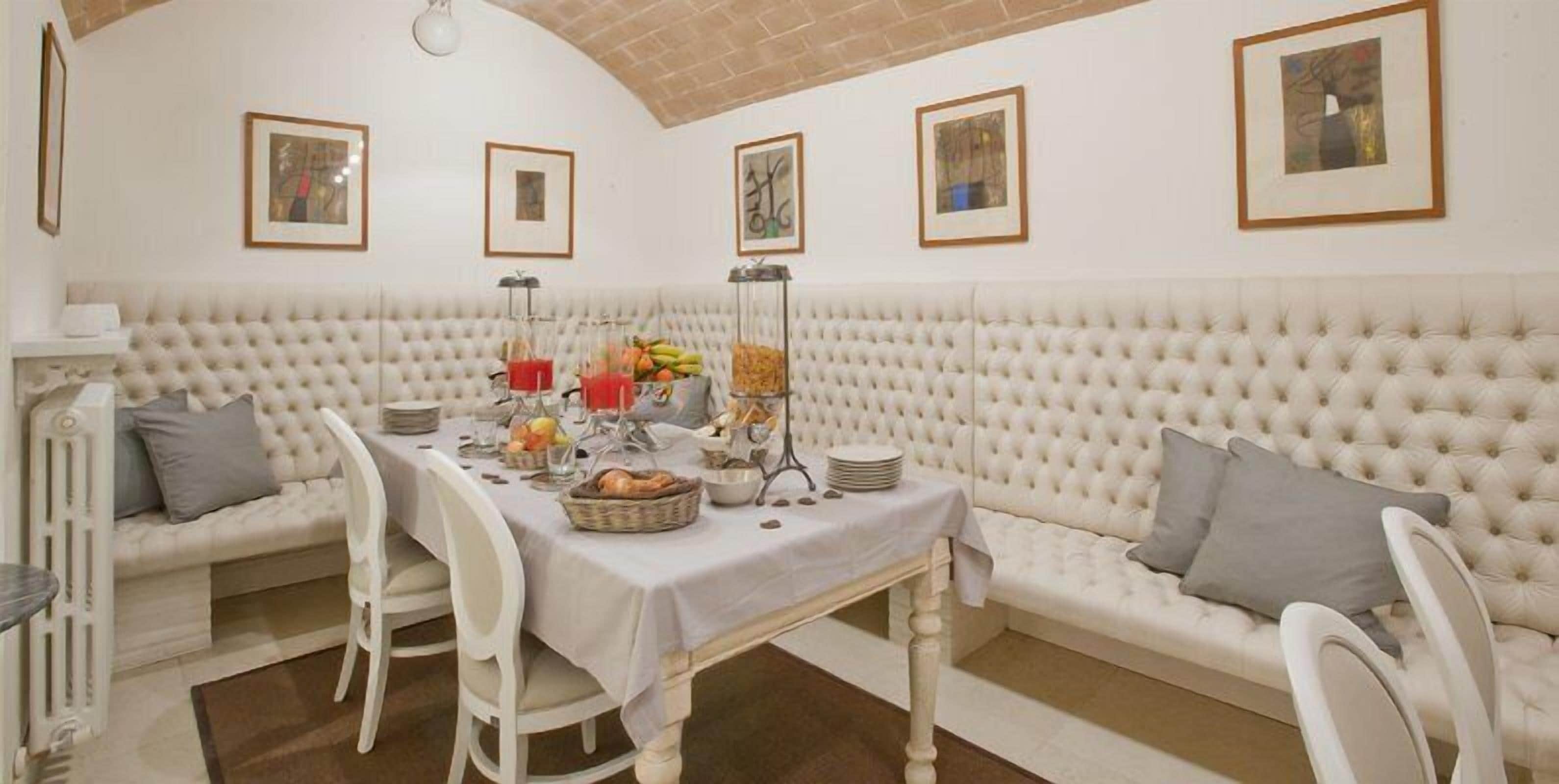 Uma sala de jantar com um sofá estofado branco, uma mesa posta com pratos, copos e frutas. Há quadros nas paredes e um radiador à esquerda.