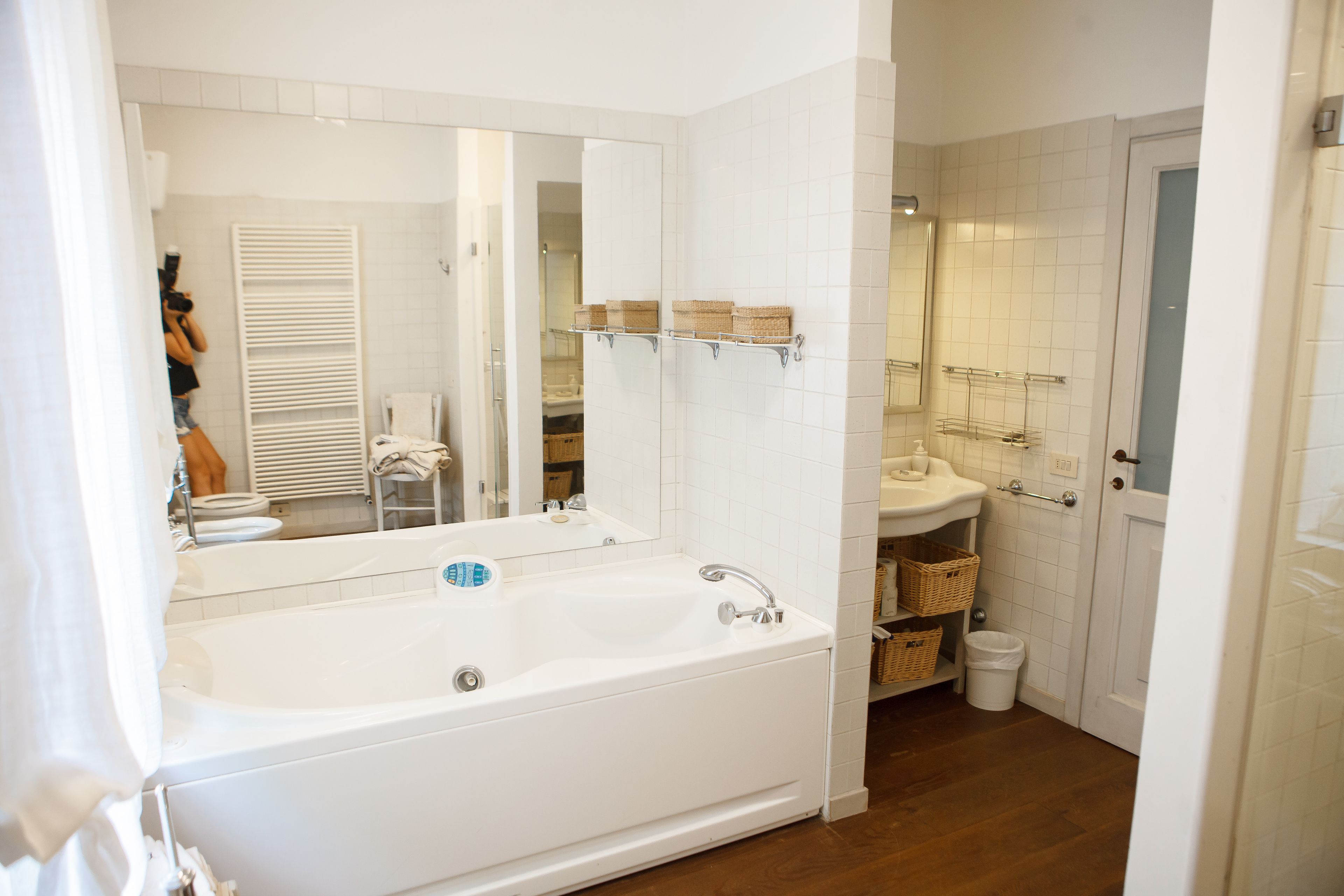 Ein modernes Badezimmer mit einer großen Badewanne, einem Waschbecken und einem Spiegel. Es gibt Regale mit Körben und eine Tür zu einem anderen Raum.